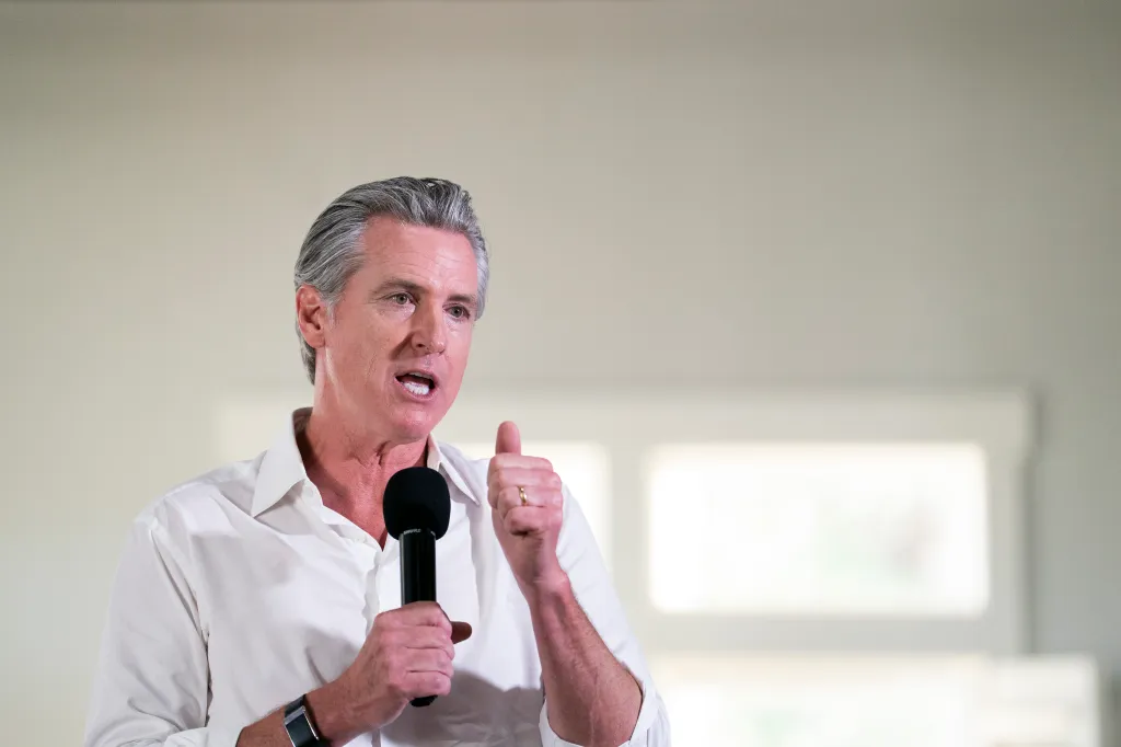 Gavin Newsom speaking at an event, holding a microphone and giving a thumbs-up.