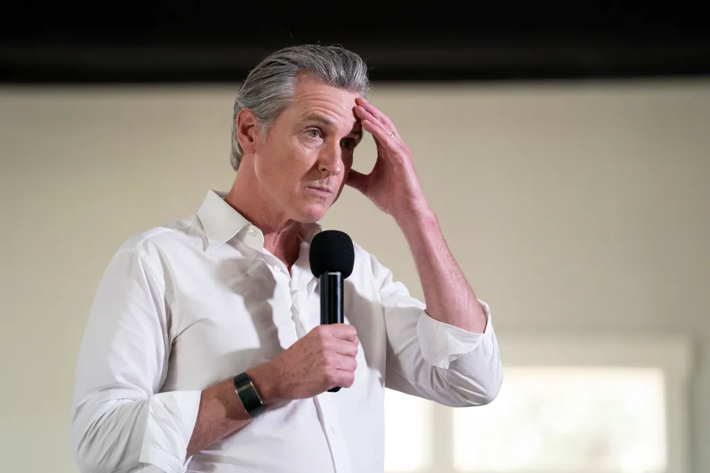 Gavin Newsom speaking into a microphone, with his left hand touching his forehead.