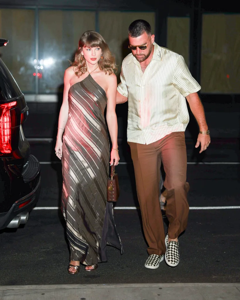 Taylor Swift in a metallic striped dress and Travis Kelce in a striped shirt and brown trousers walk together at night.
