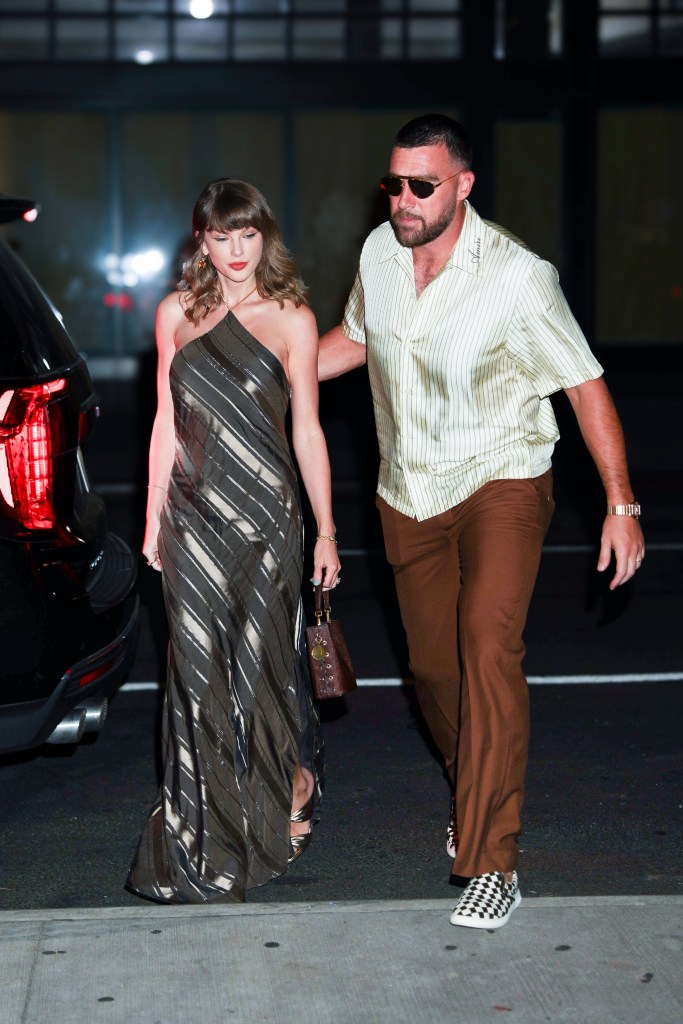 Taylor Swift in a metallic striped dress and Travis Kelce in a striped shirt and brown trousers walking outdoors at night.