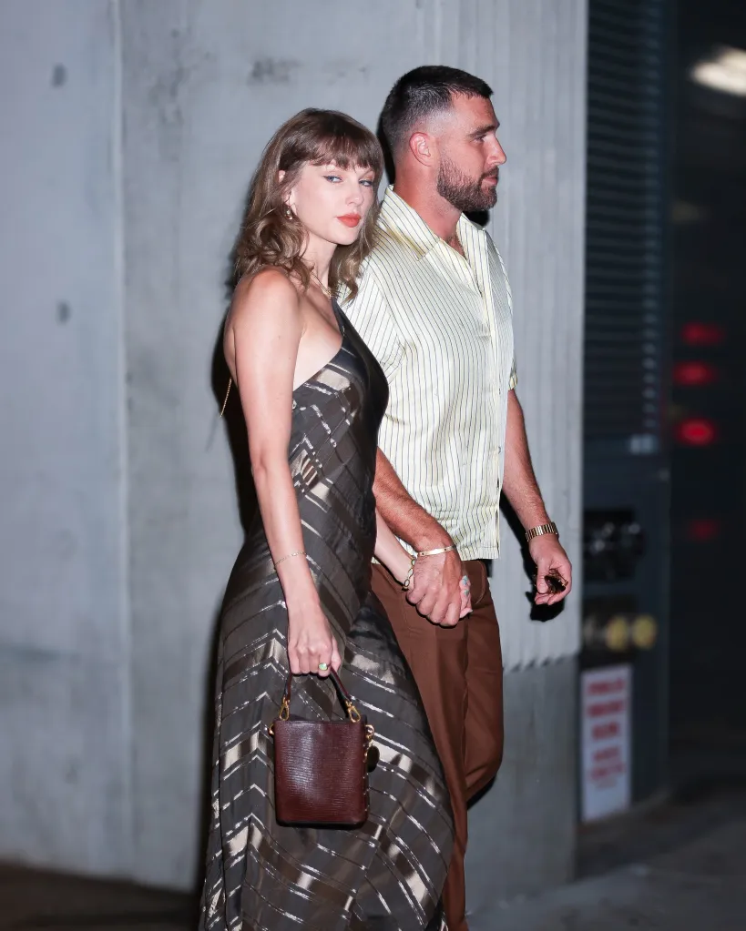 Taylor Swift and Travis Kelce walking hand-in-hand while Swift carries a small purse and wears a metallic striped dress and Kelce wears a striped short-sleeve shirt and brown trousers.
