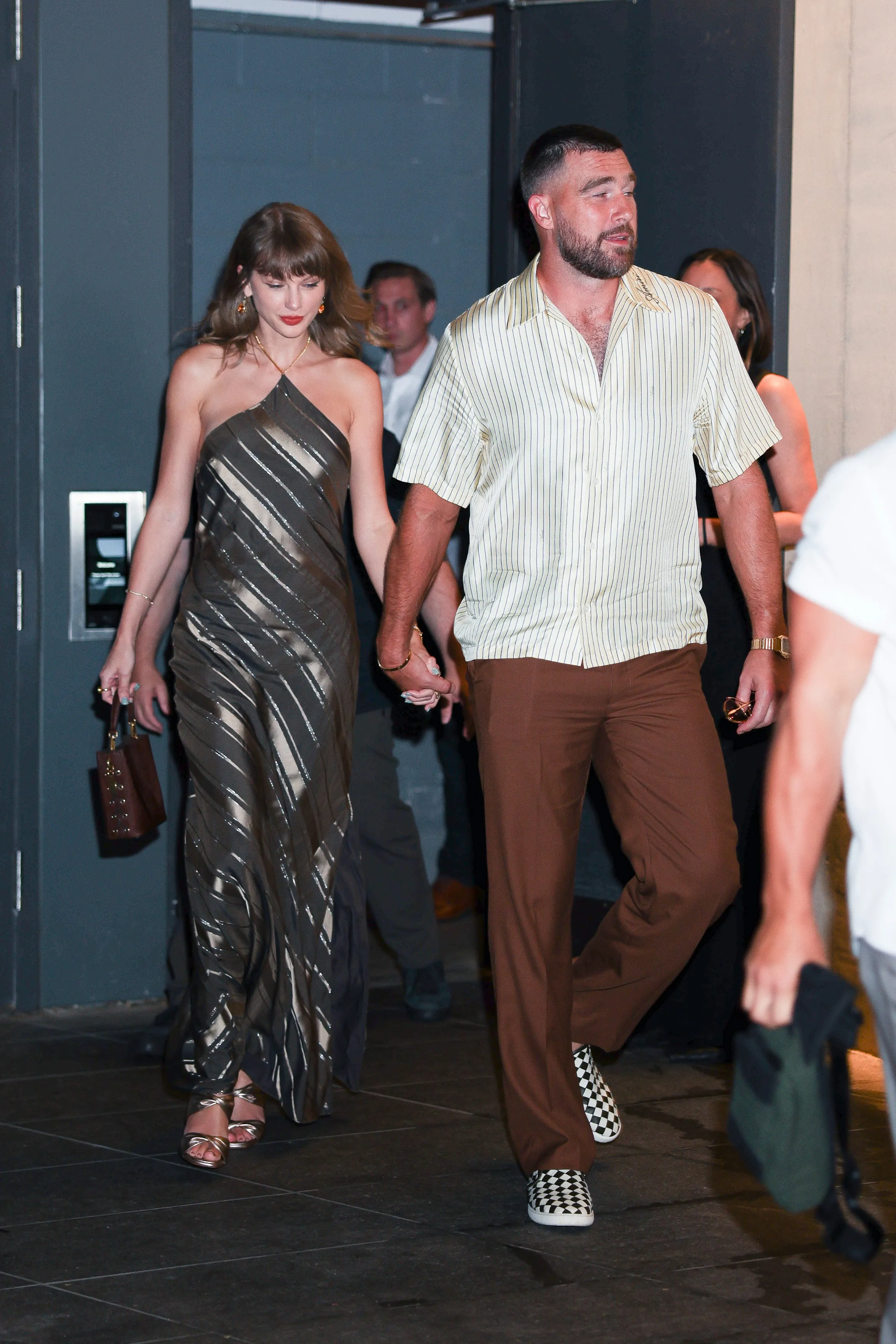 Taylor Swift in a metallic striped dress and Travis Kelce in a striped shirt and brown trousers walking hand-in-hand to dinner.
