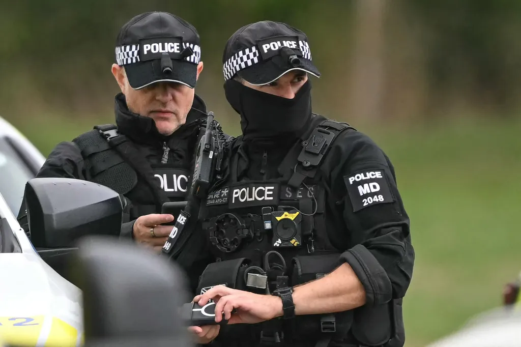 Two police officers, one partially obscured by a car, wearing black uniforms, caps, and tactical gear at an incident site.