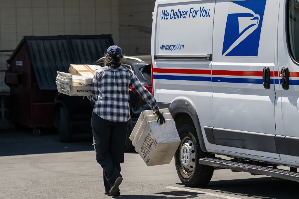 A United States Postal Service worker carries mail containers and packages.