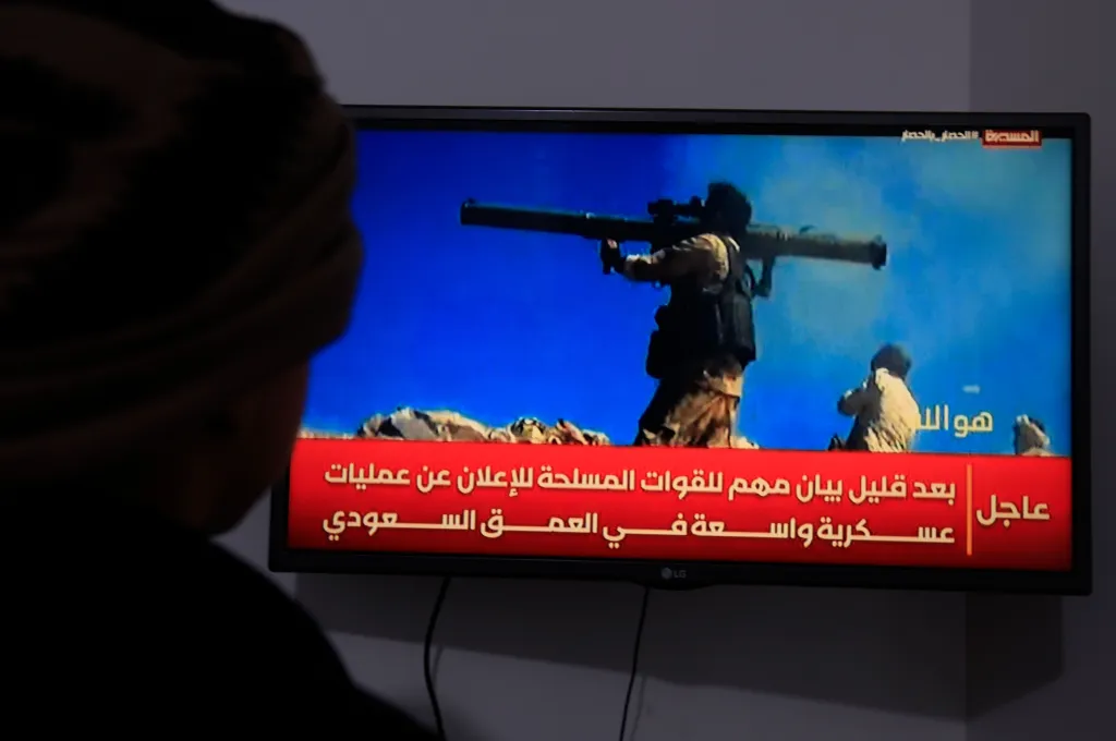 A person watches a TV broadcast about the Saudi-Houthi conflict, showing a soldier with a rocket launcher and Arabic text.