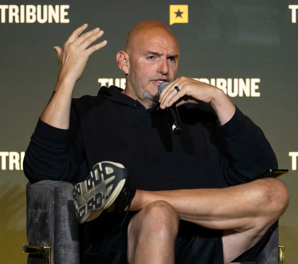 John Fetterman speaking into a microphone, gesturing with his left hand, and wearing a black hoodie and shorts at the Texas Tribune Festival.