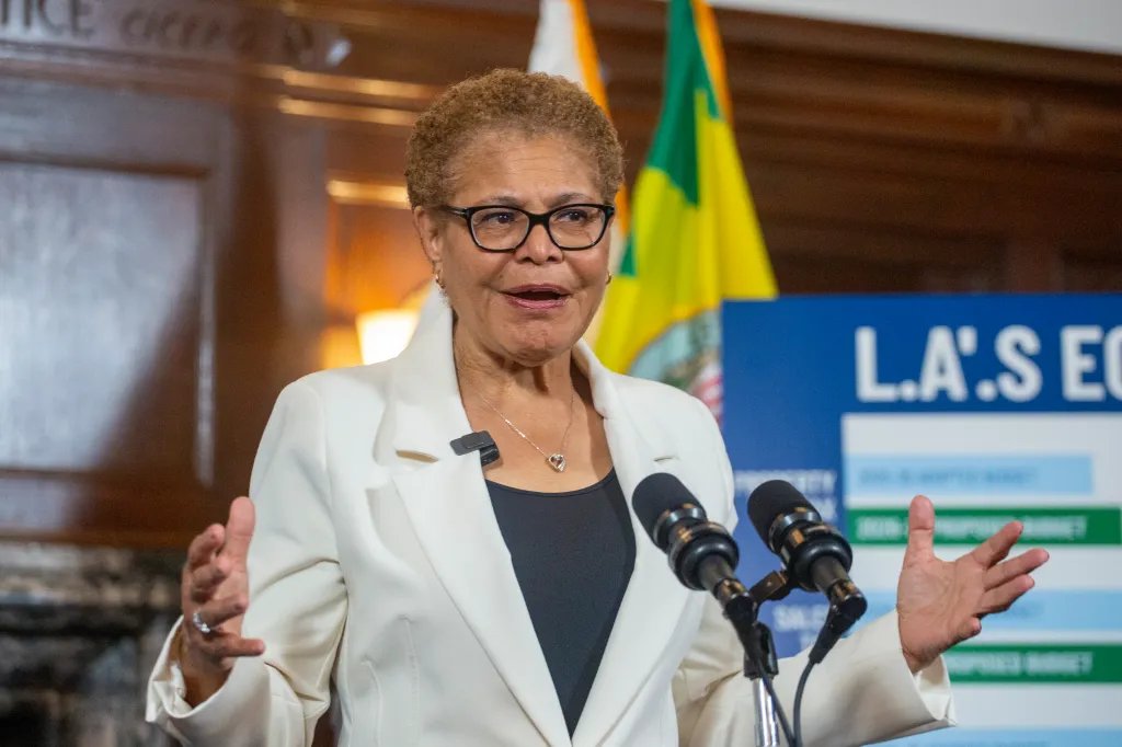 Mayor Karen Bass speaking at a podium with microphones and a display board behind her.