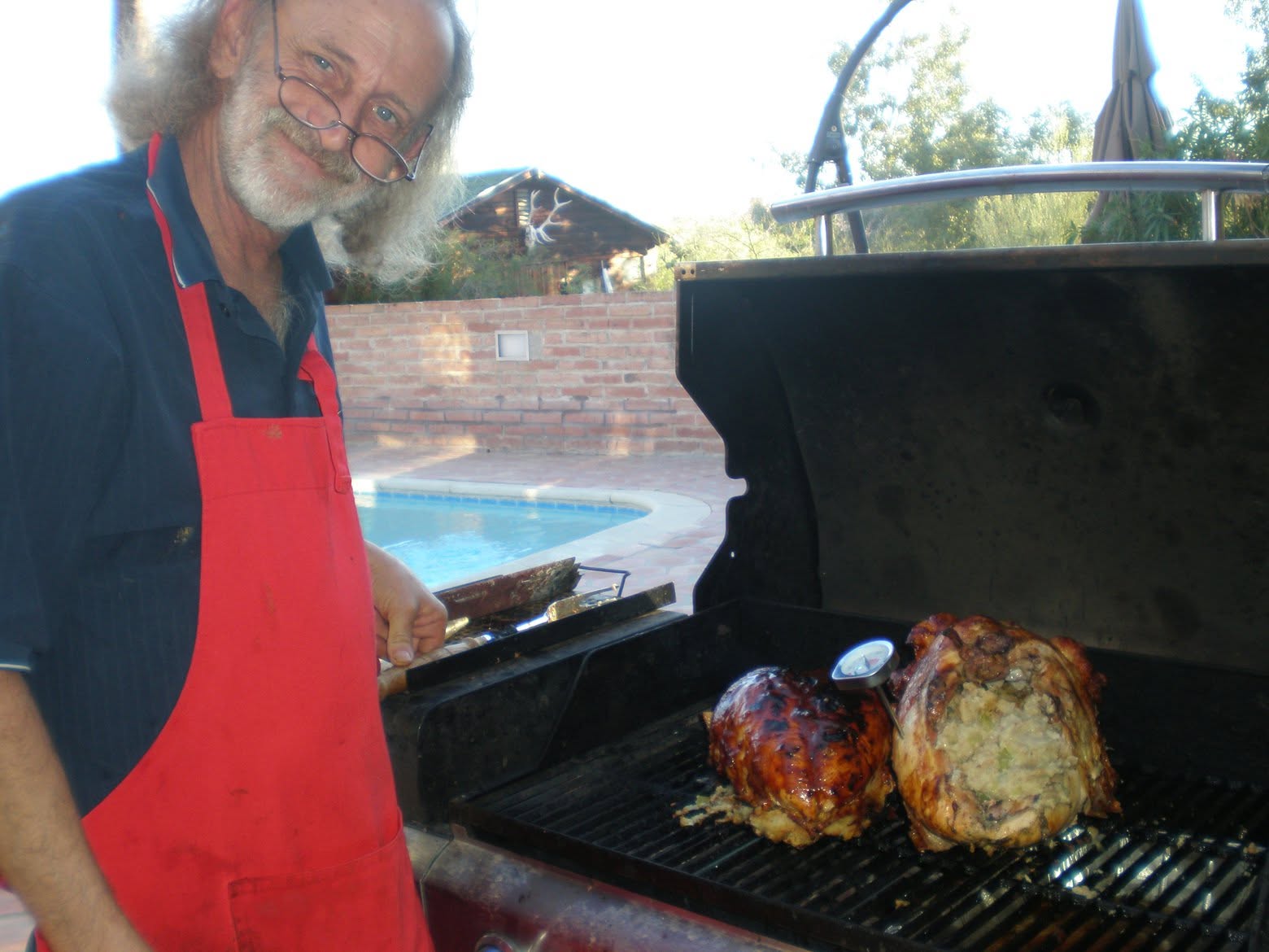 Christopher Nicks, wearing a red apron, barbecues two stuffed chickens by a swimming pool.