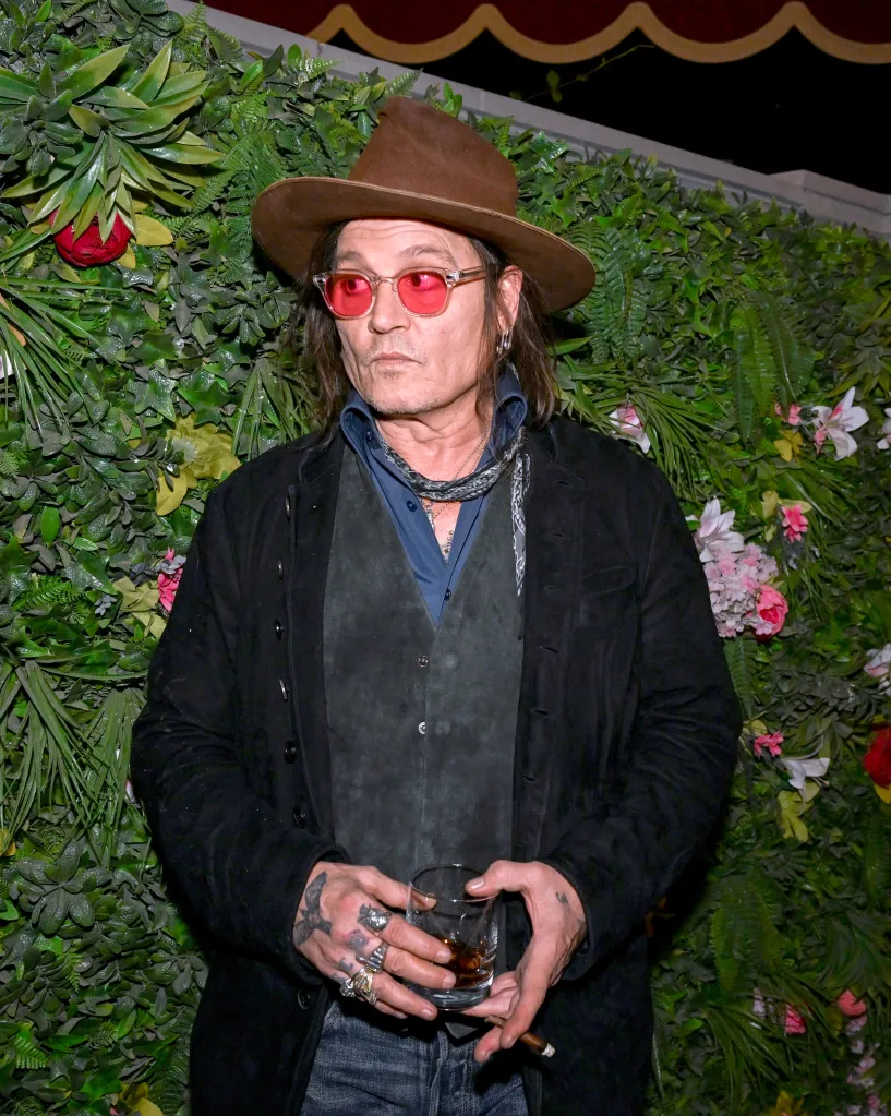 Johnny Depp in a brown hat and red sunglasses, holding a drink and cigar, standing against a green leafy backdrop.