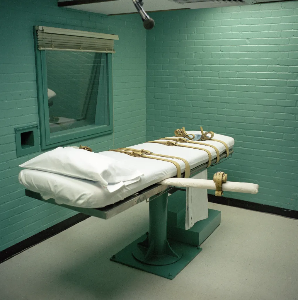 A gurney with straps in a teal-green execution chamber with a window looking into another room.