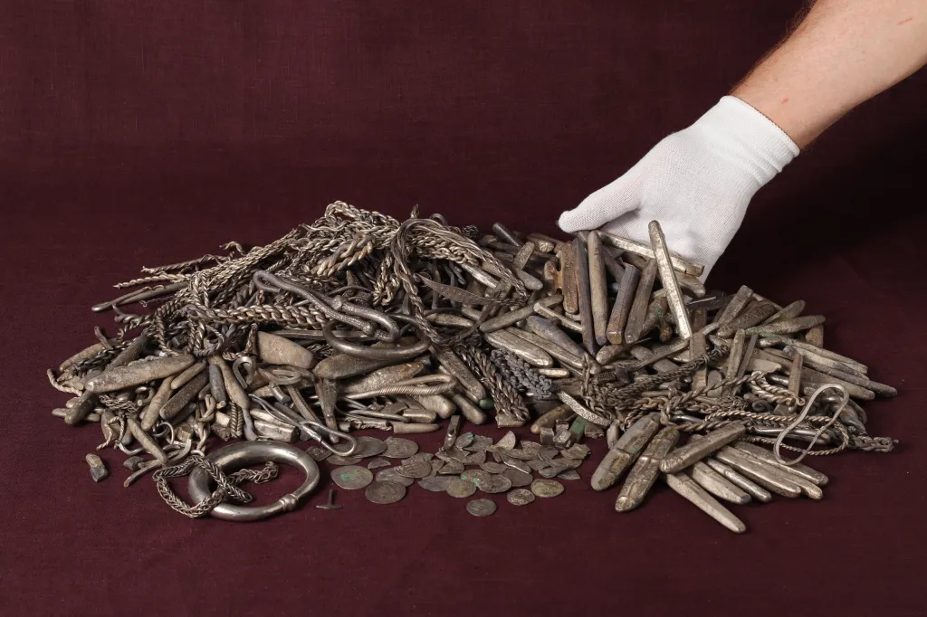 A gloved hand rests on a large pile of ancient silver coins, bracelets, and bars, part of Denmark's largest Viking silver hoard.
