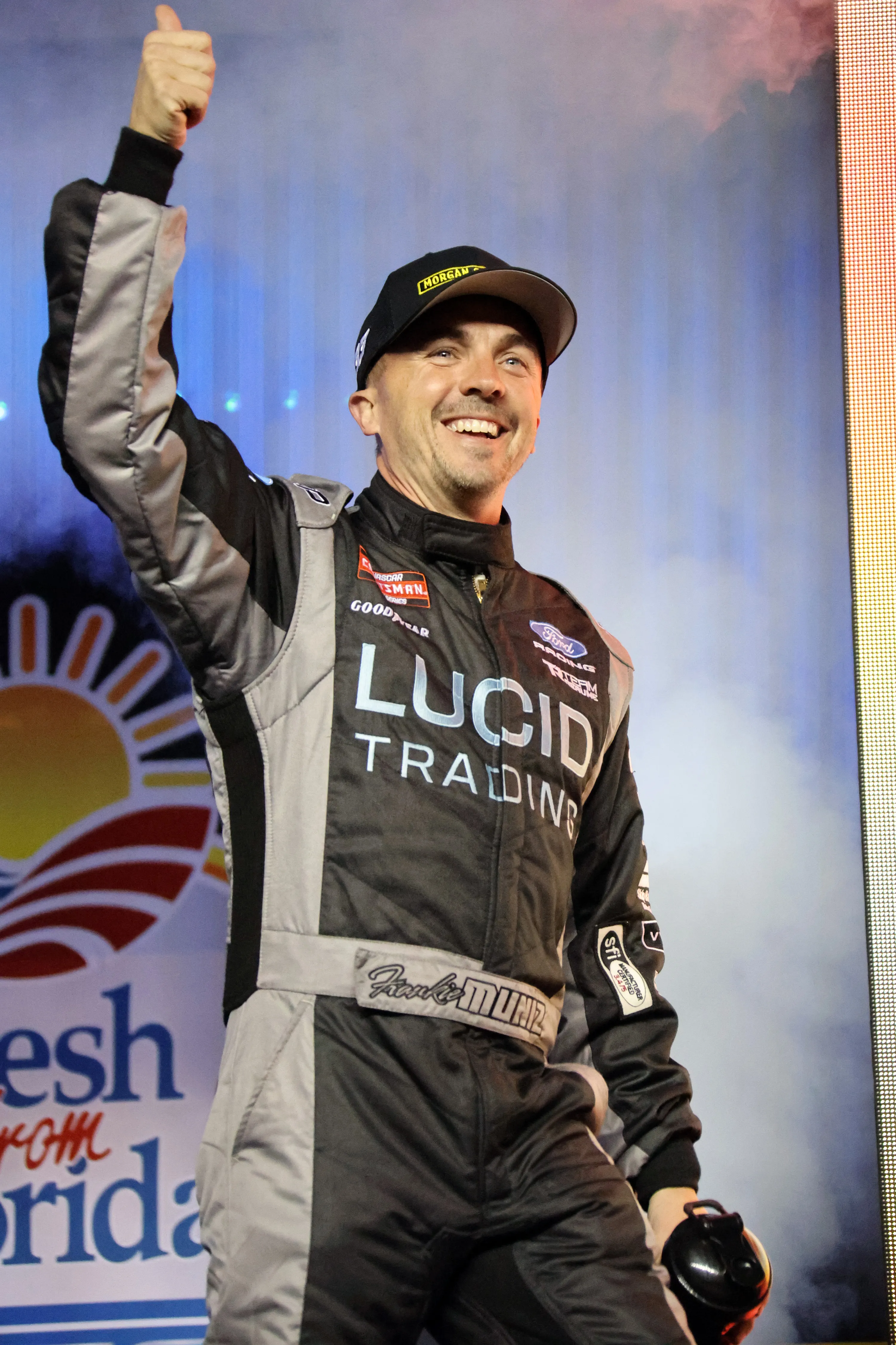 Frankie Muniz in a black and grey racing suit and cap, holding his helmet and giving a thumbs up.