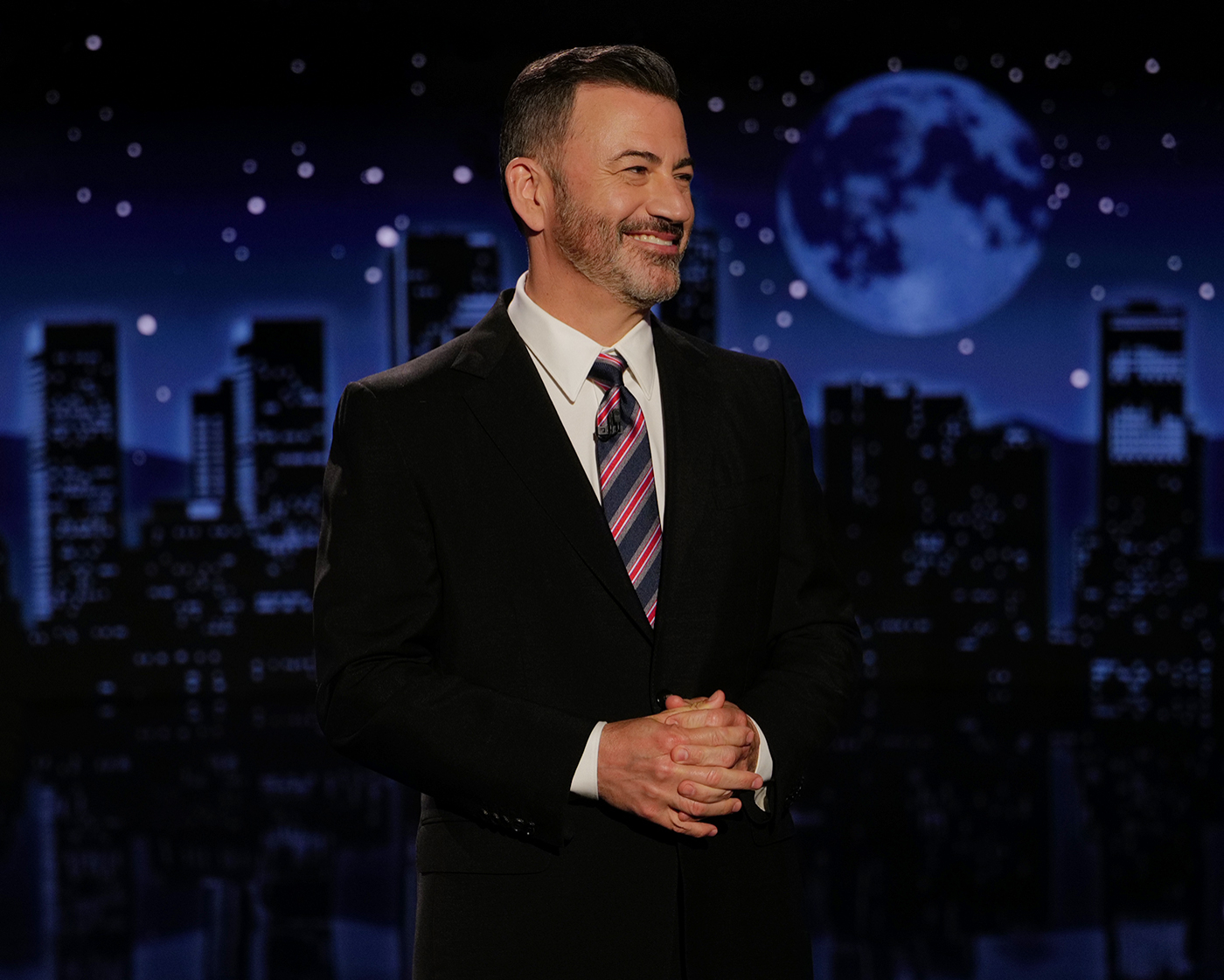 Jimmy Kimmel smiling on stage in a suit and tie, with a cityscape and full moon in the background.