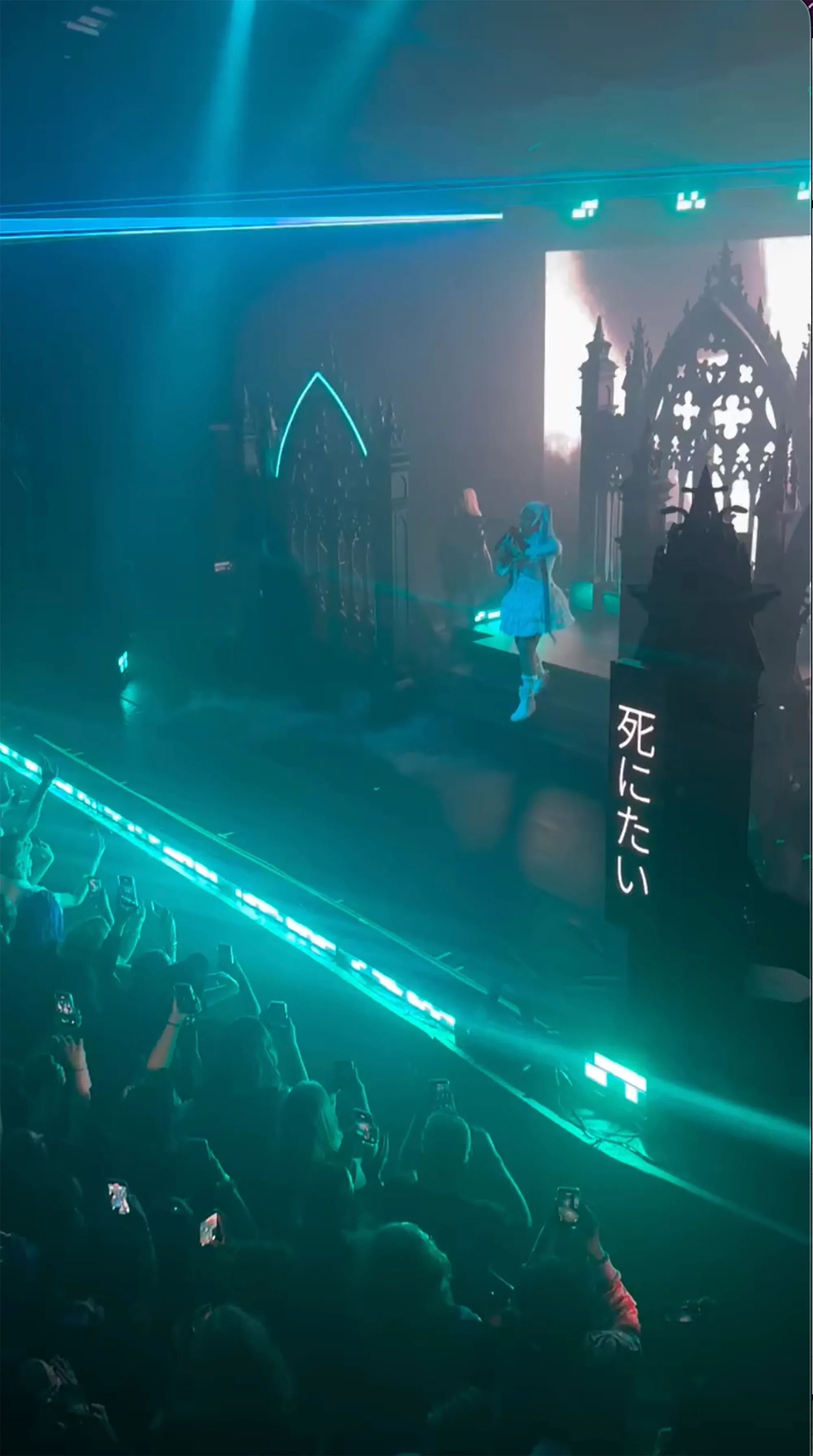 North West performing on stage in a light blue dress with gothic-style set pieces and a crowd recording on their phones.