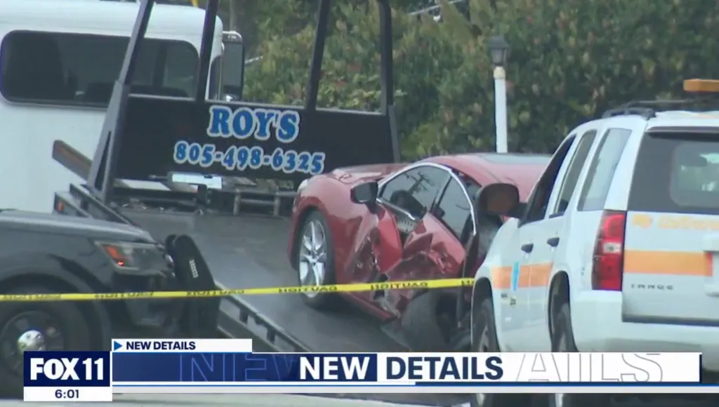 Damaged red car on a tow truck, with police vehicles and caution tape visible.