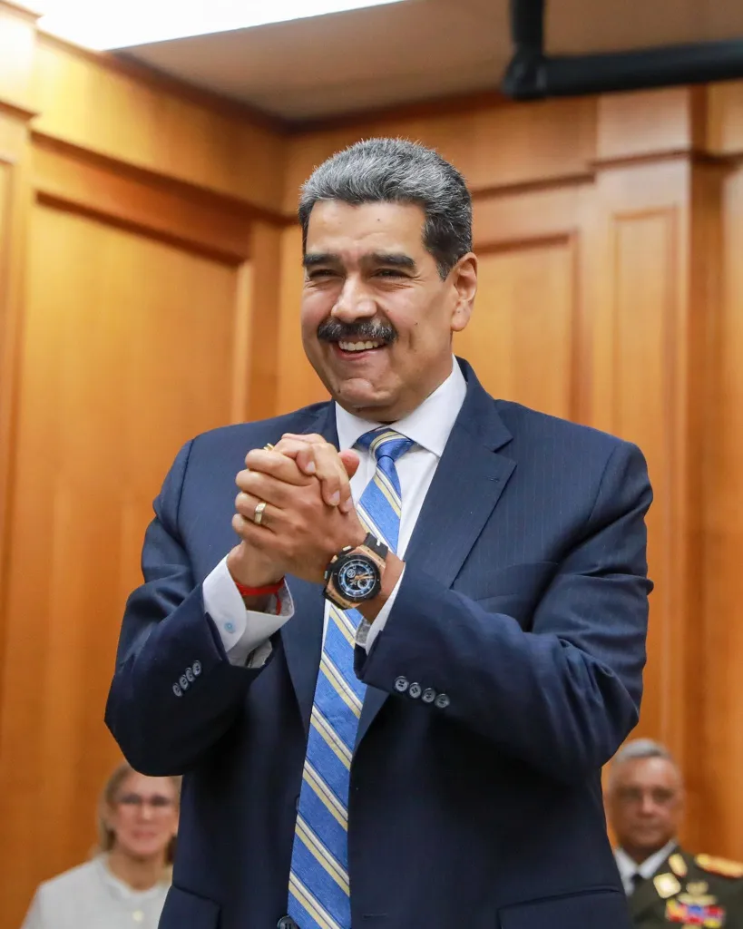 Venezuelan President Nicolas Maduro in a dark suit and blue striped tie, clasping his hands and smiling.