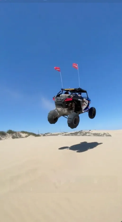 A dune buggy with two red flags attached is airborne over sand dunes against a bright blue sky.