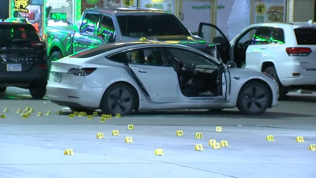 A white Tesla riddled with bullet holes and its doors ajar, surrounded by yellow evidence markers on the ground.