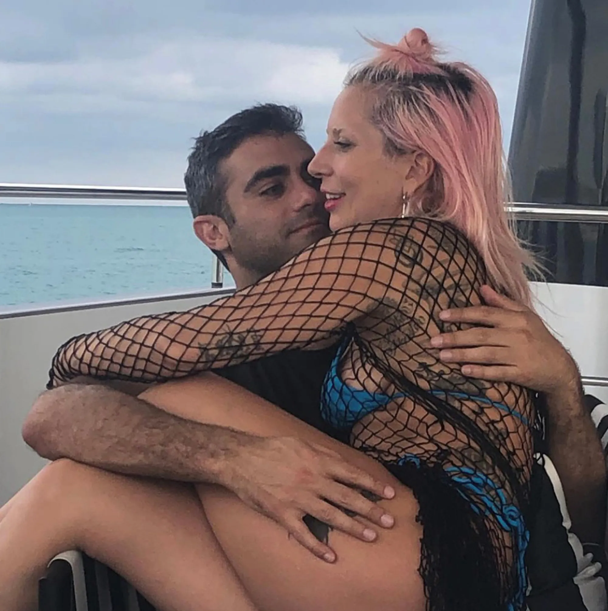 A man in a black shirt hugs a woman with pink hair, wearing a fishnet top and blue bikini, on a boat.