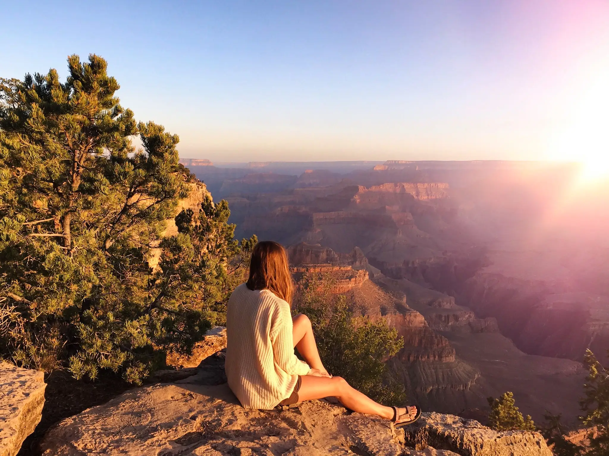 Emily, wearing a cream sweater, shorts, and sandals, sits on a lookout at the Grand Canyon at sunset.