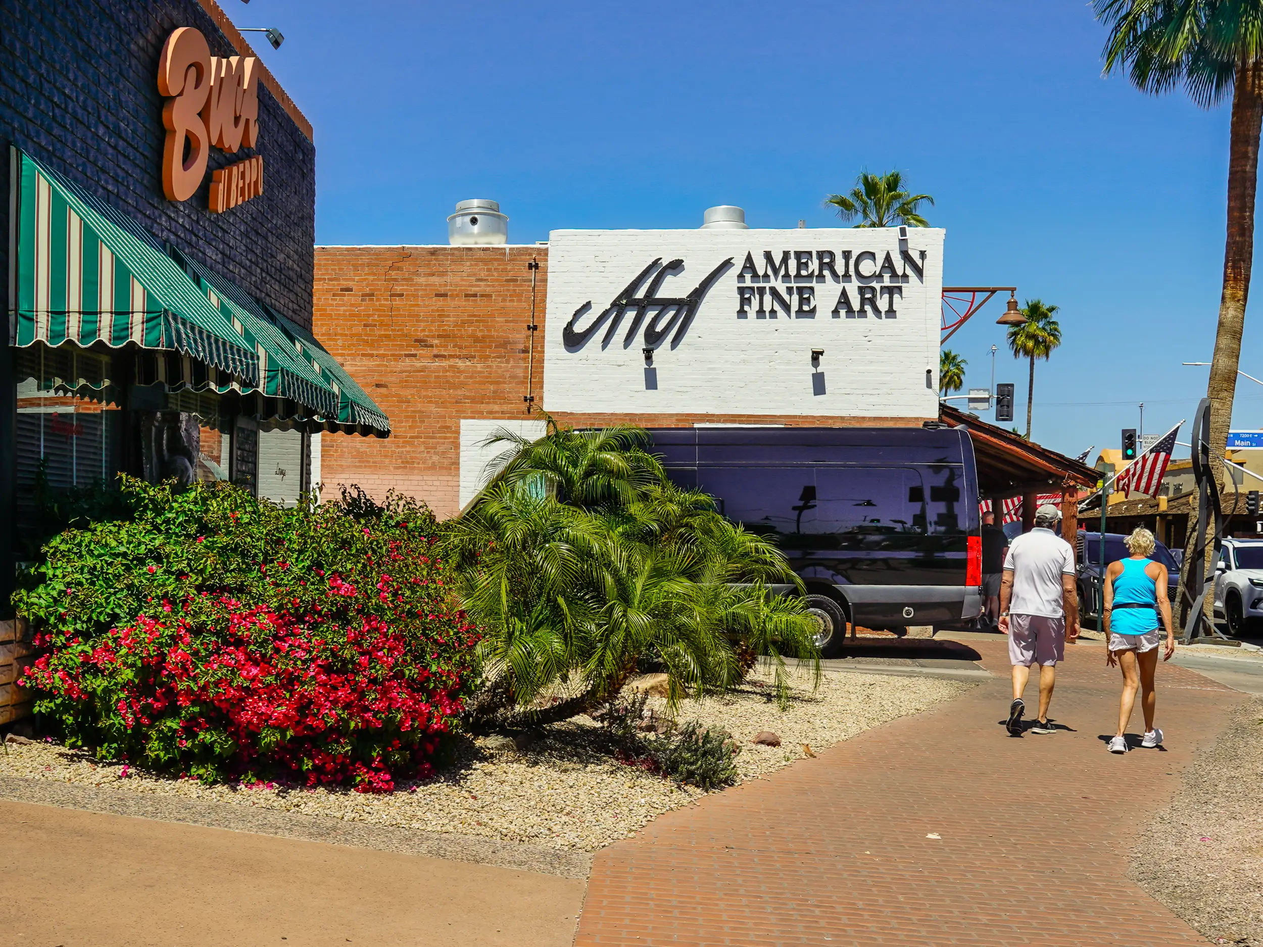 A brick path with storefronts and bushes on the left and a tree on the right on a clear day with blue skies