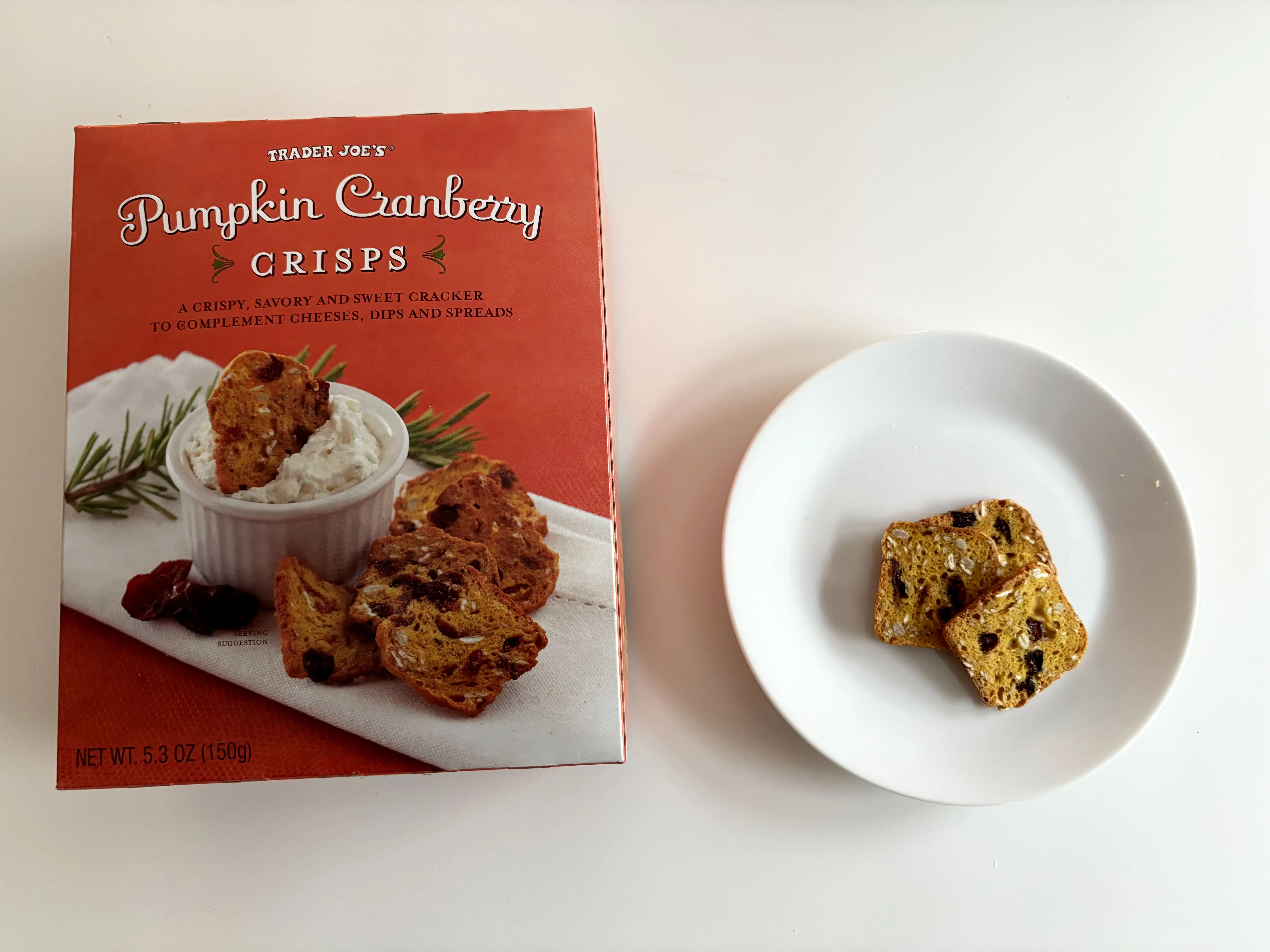 A box of Trader Joe's pumpkin-cranberry crisps next to a white plate with crackers on it.