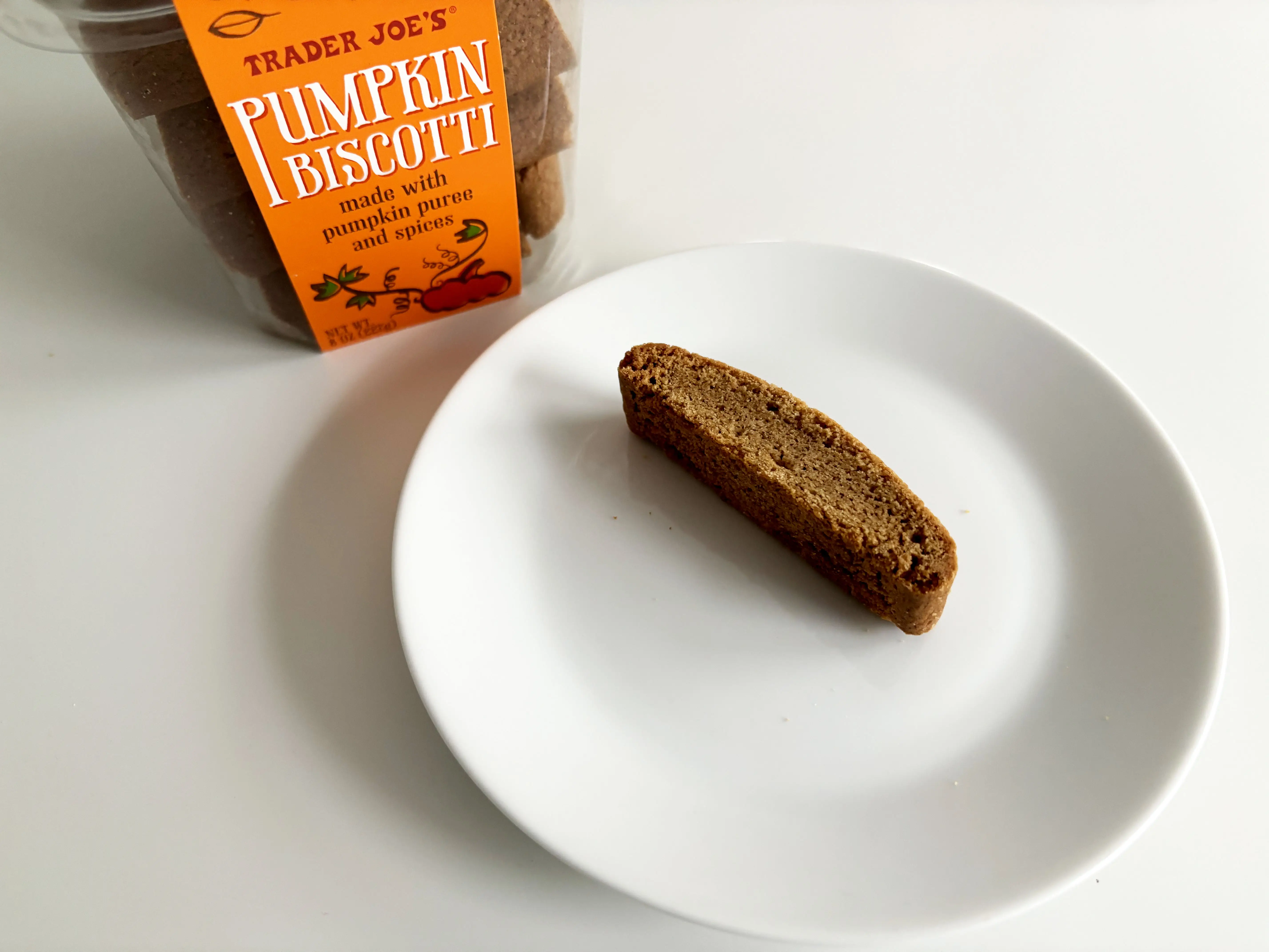 A box of Trader Joe's pumpkin biscotti next to a white plate with a cookie on it.