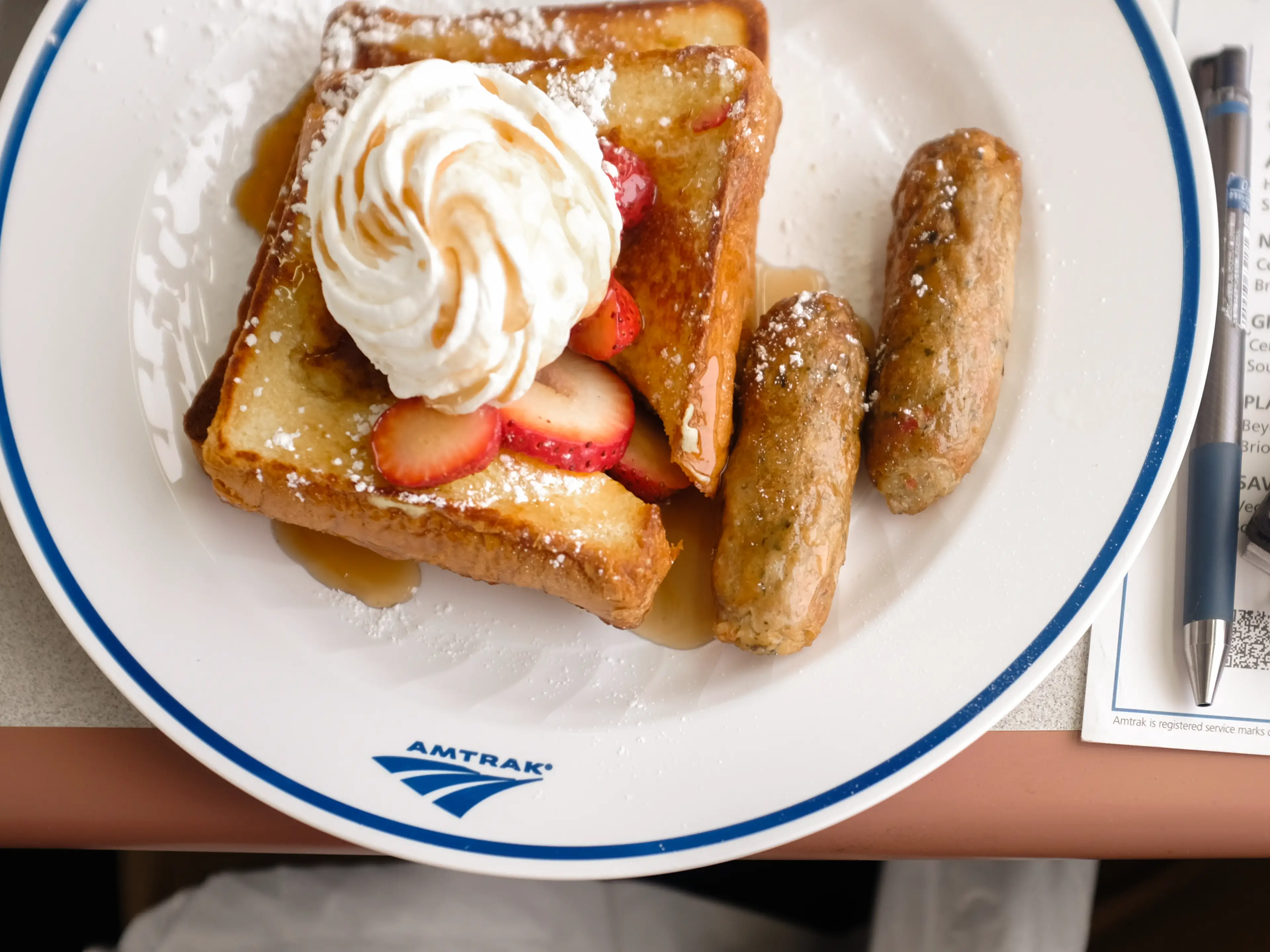 A plate of french toast with sausage, syrup, whipped cream, and strawberries.