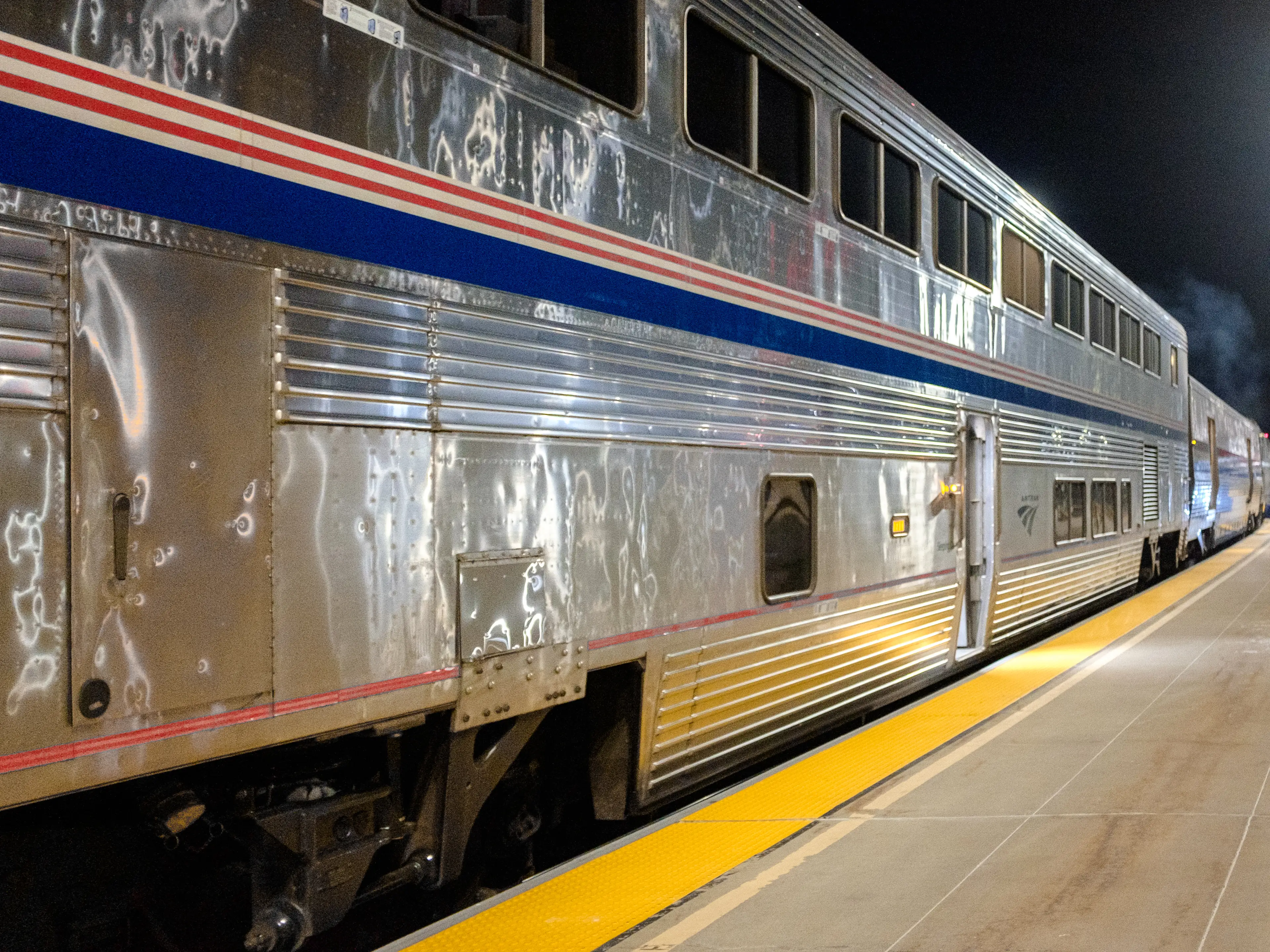 A gray train with red and blue stripes parked at a platform at night
