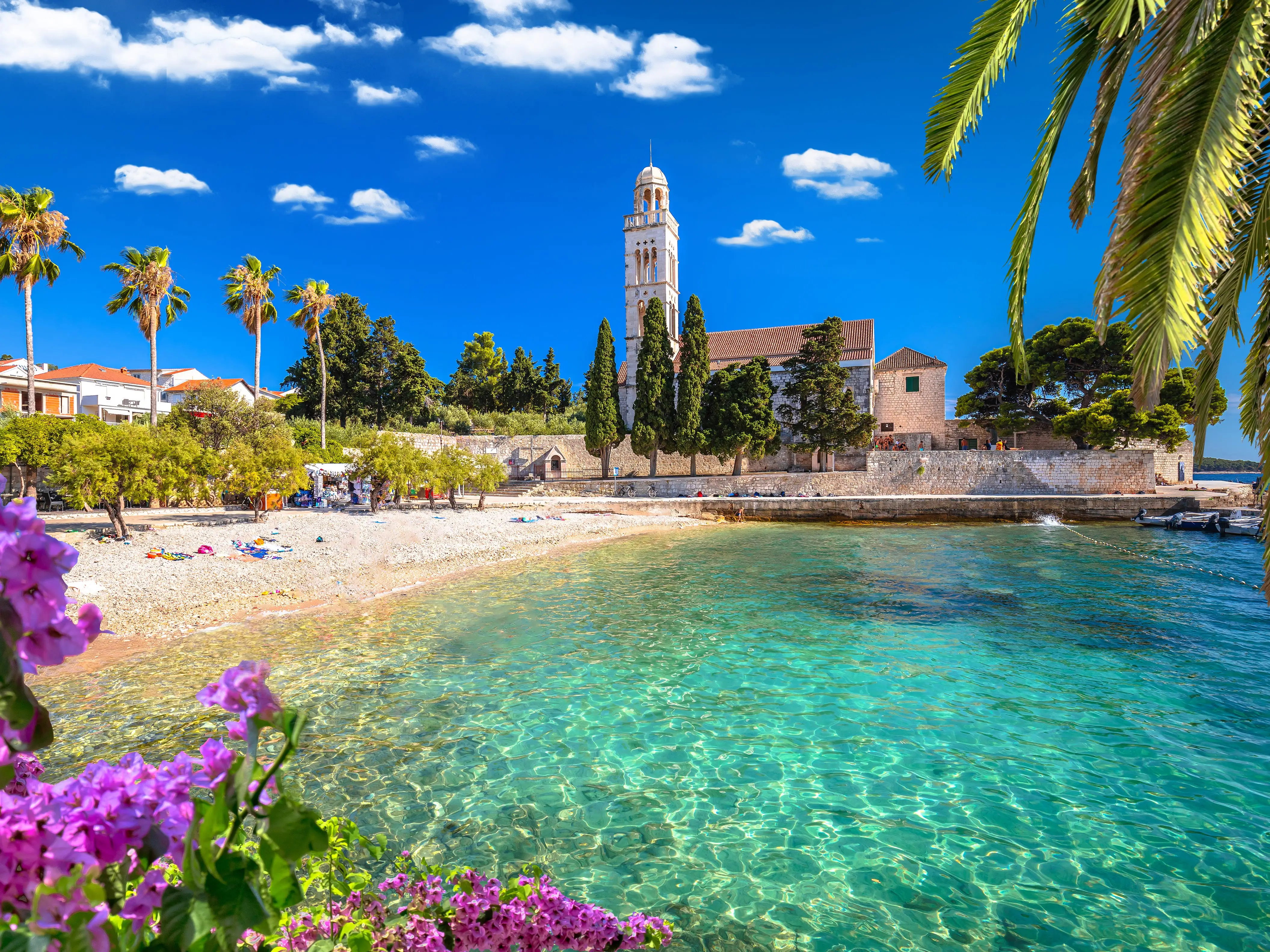 A beach with turquoise waters, a palm tree, and purple flowers in the foreground, and a church and buildings in the background.