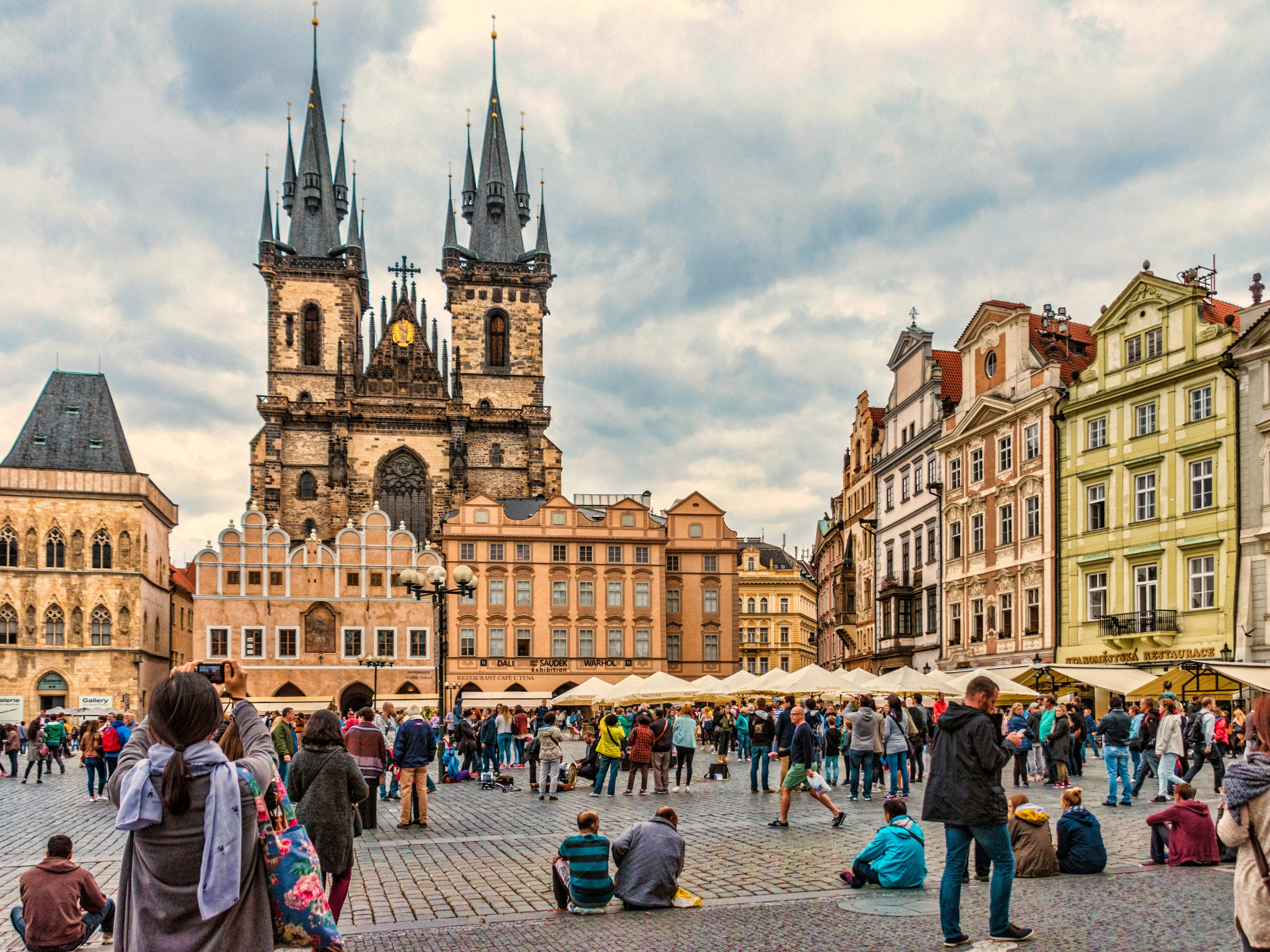 Tourists gather in a courtyard in Prague, with colorful buildings and a large church.