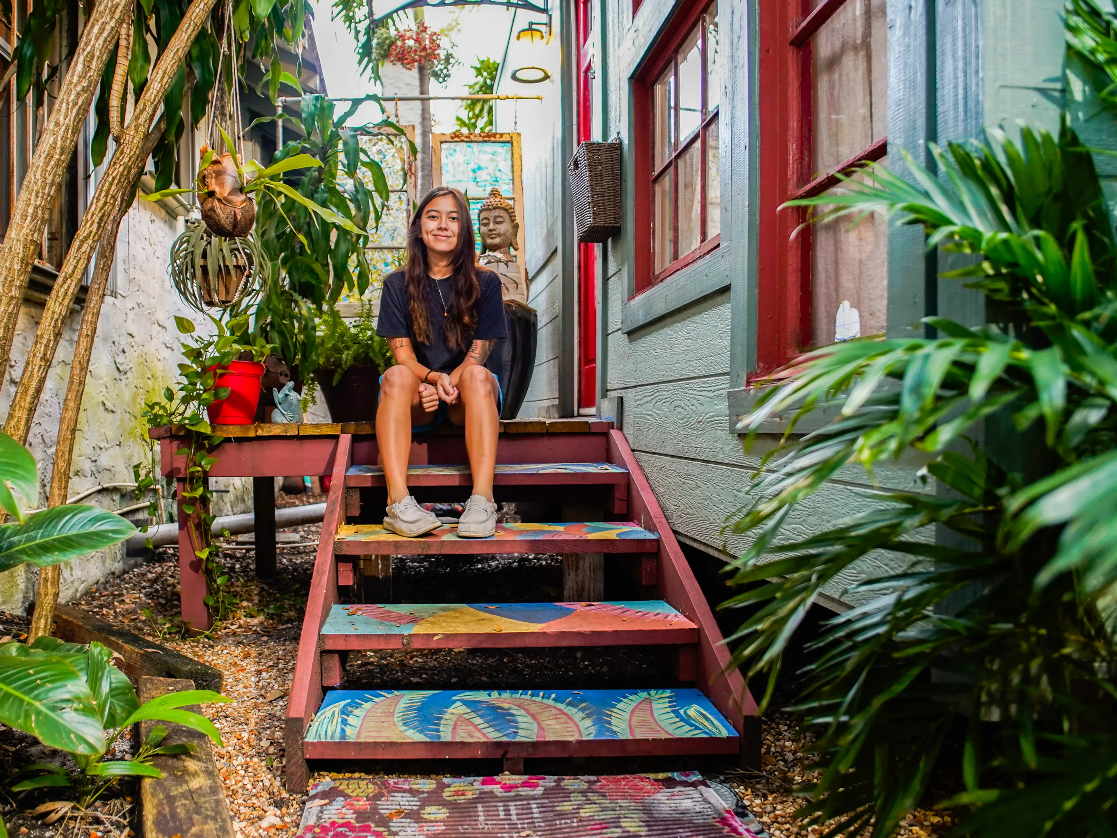 The author sits on a colorful porch surrounded by greenery outside of a tiny home on the right