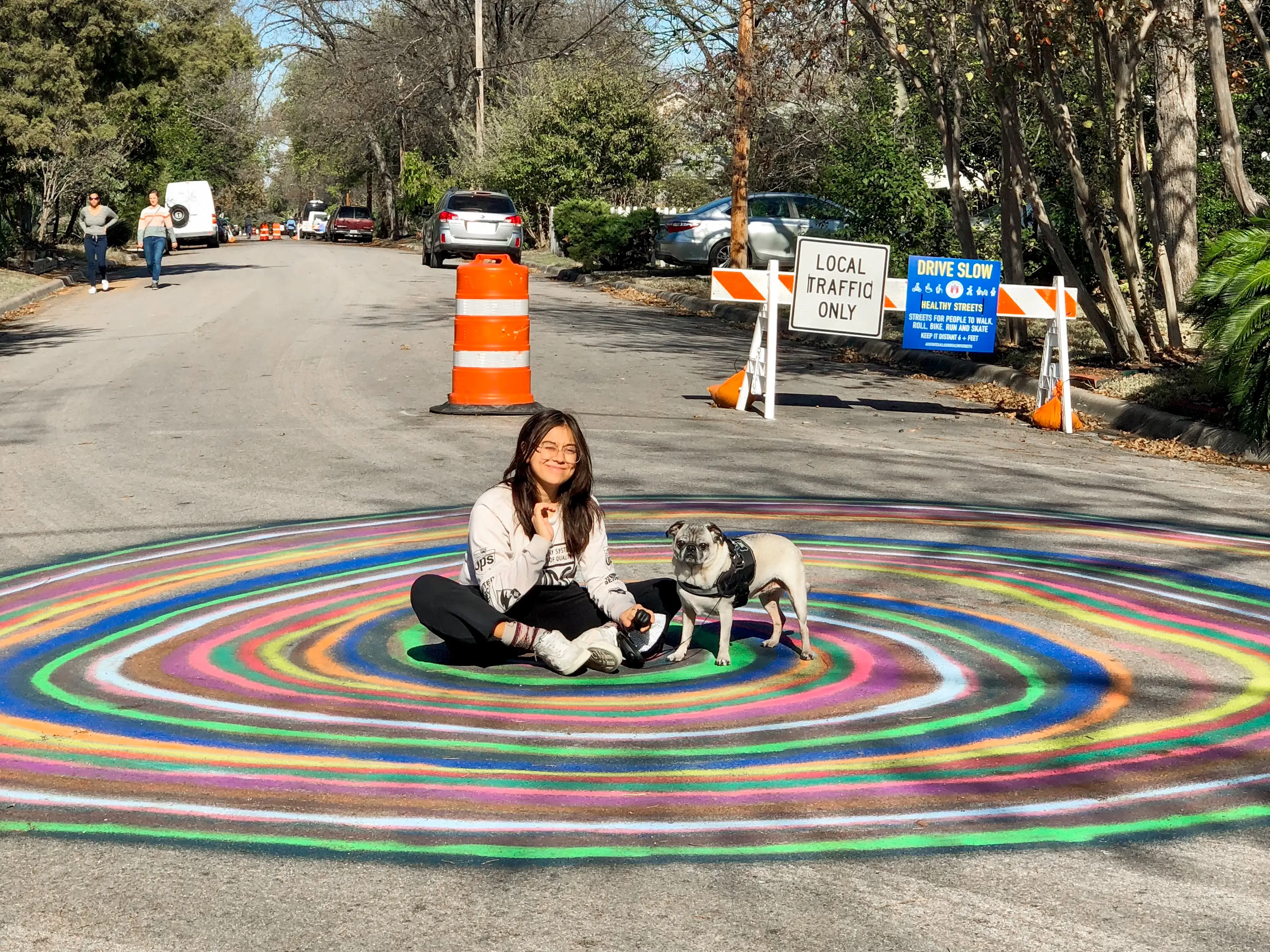 A woman sits with a pug in the center of a colorful spiral chalk drawing on a street.