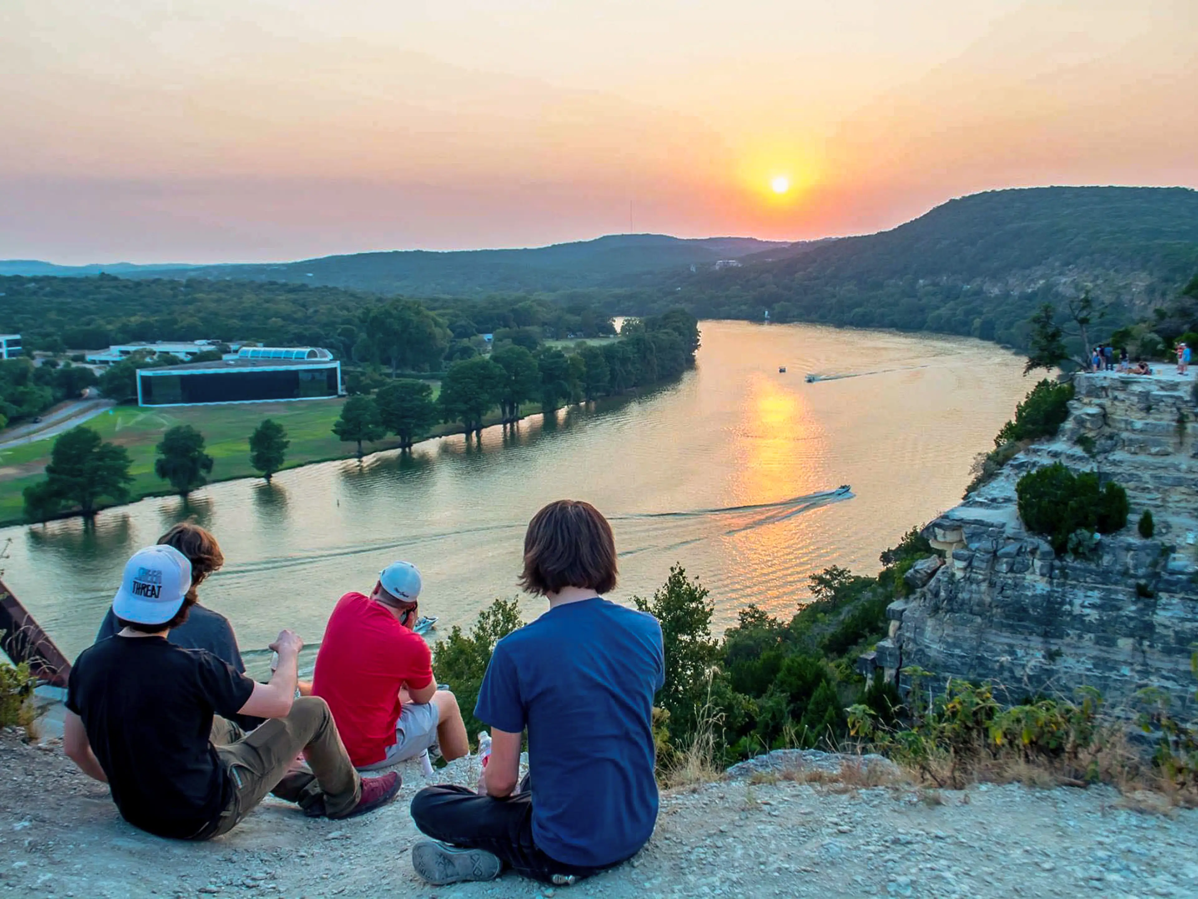A group of friends sit on the side of a cliff with rivers and hills in the background at sunset