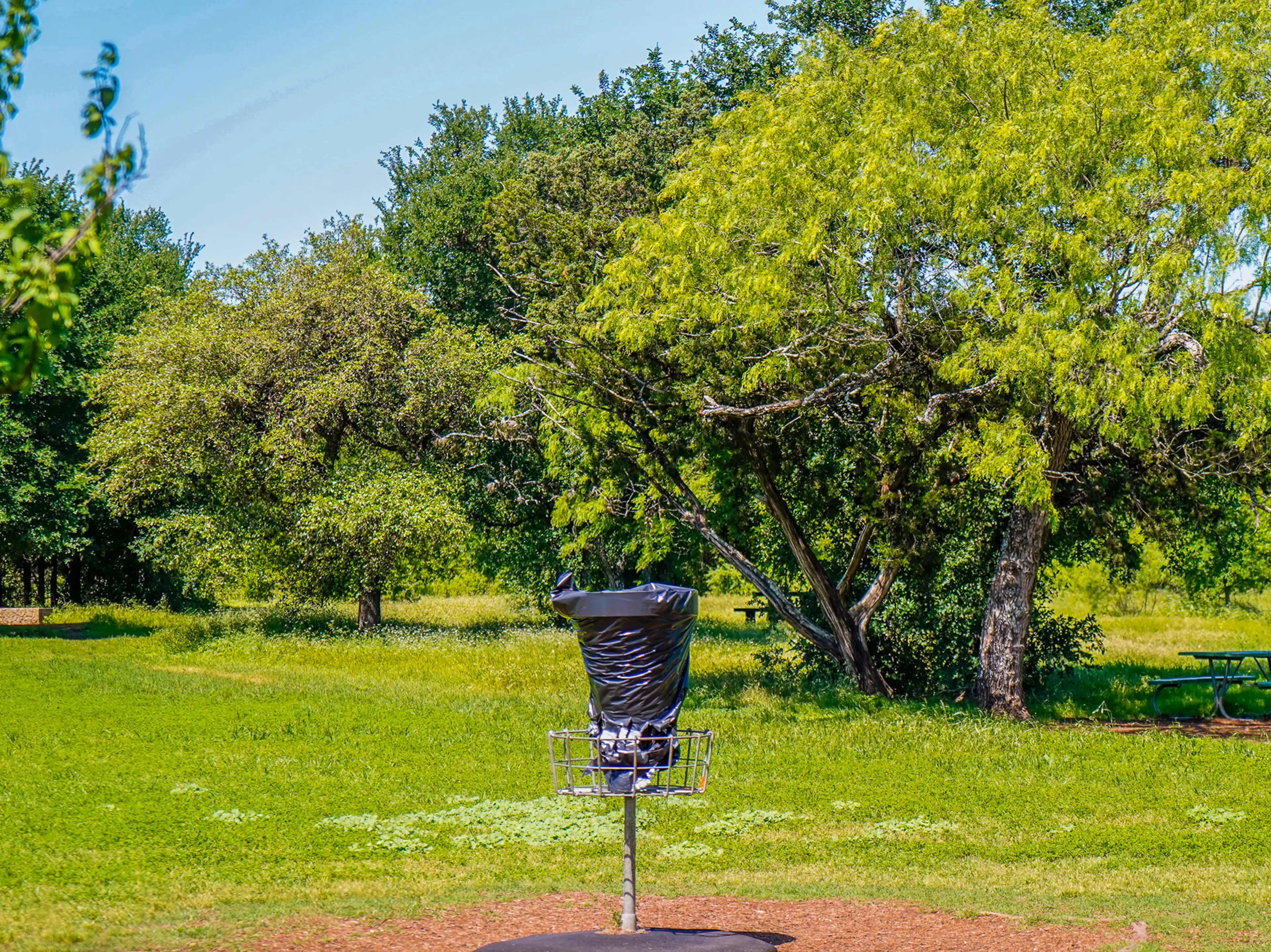 A covered disc golf basket in a green field with trees behind