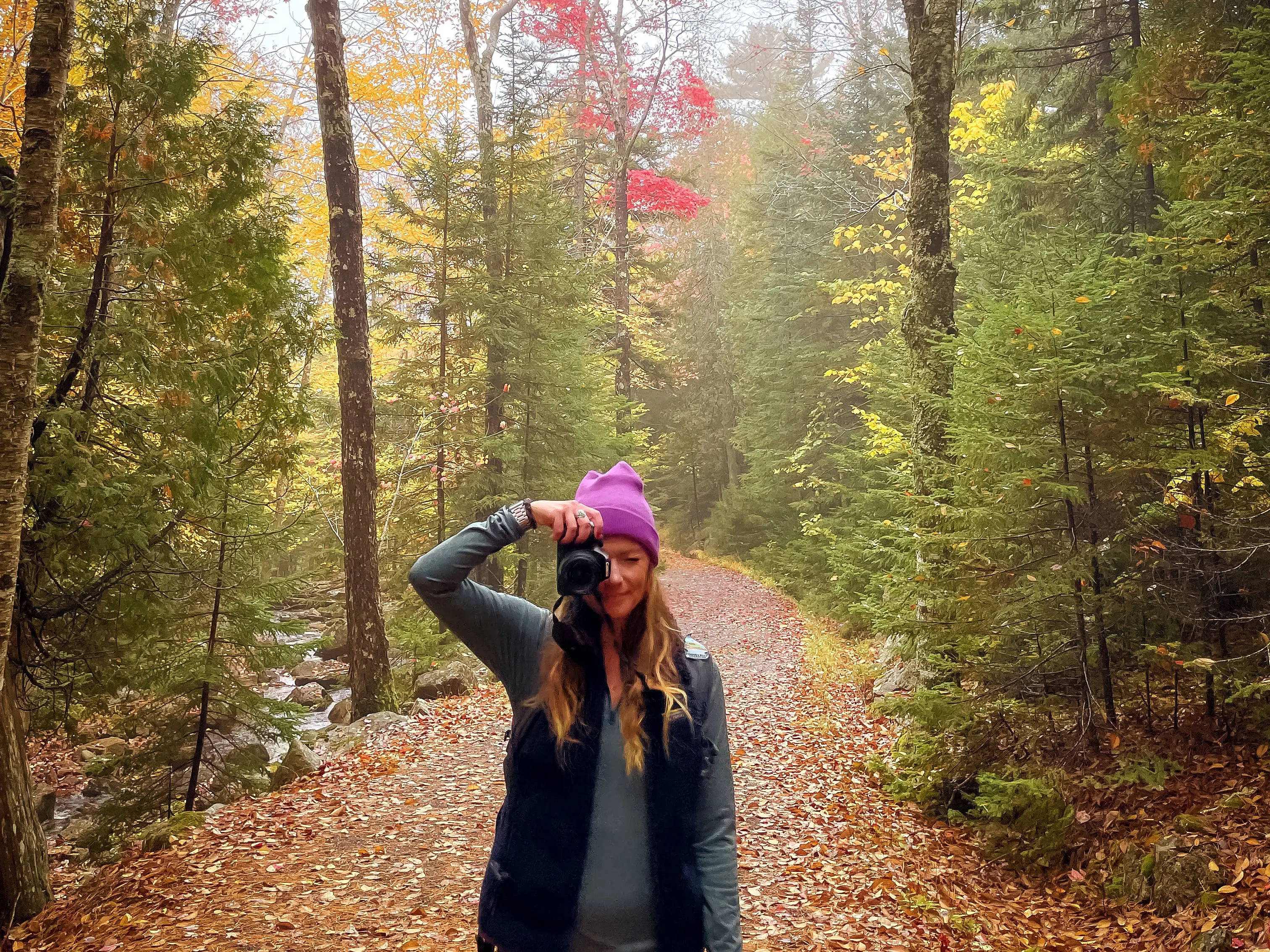 Emily holds up a camera while surrounded by colorful trees and fallen autumn leaves.