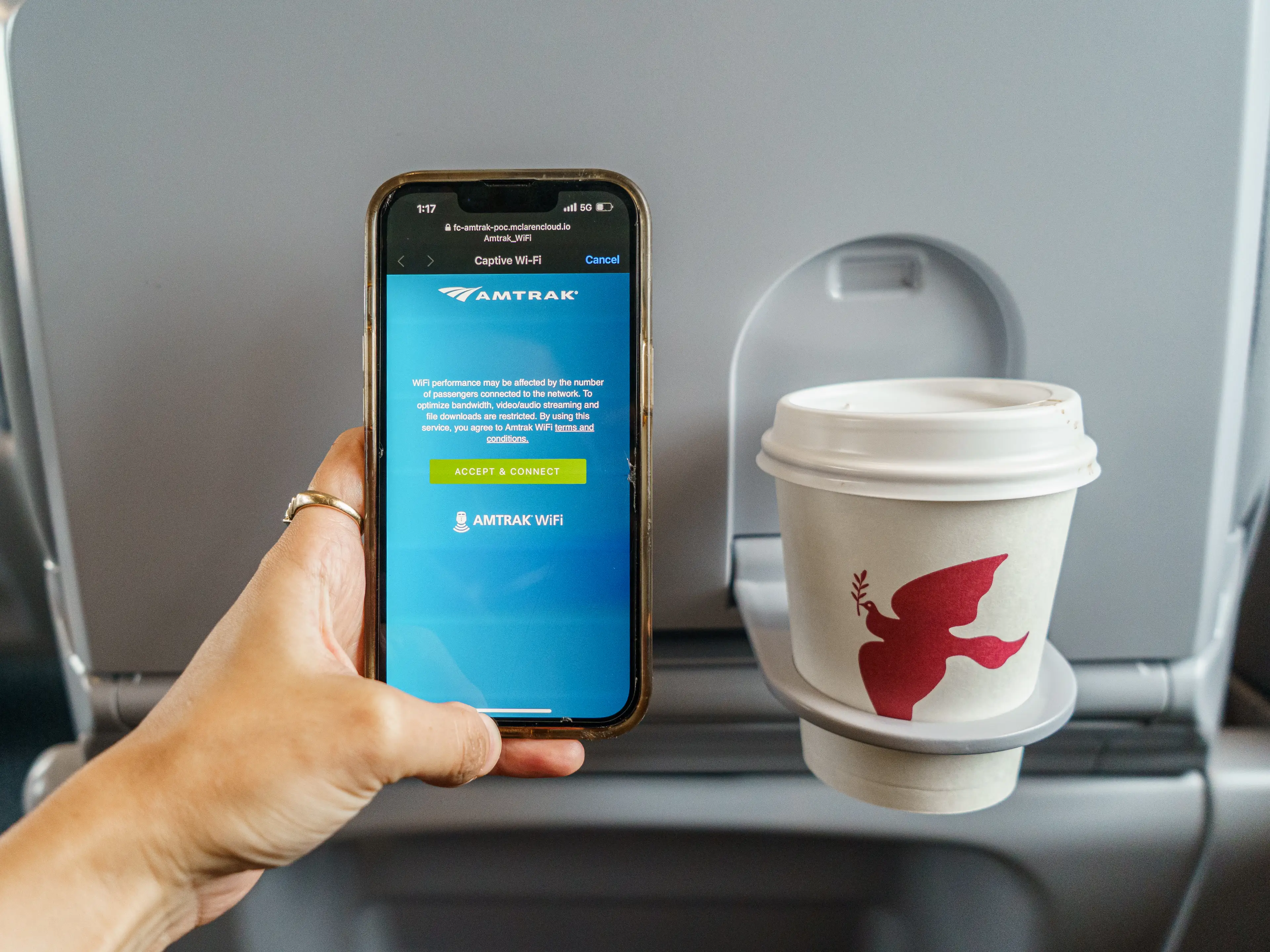 A hand holds a phone next to a coffee cup in front of a train seat back