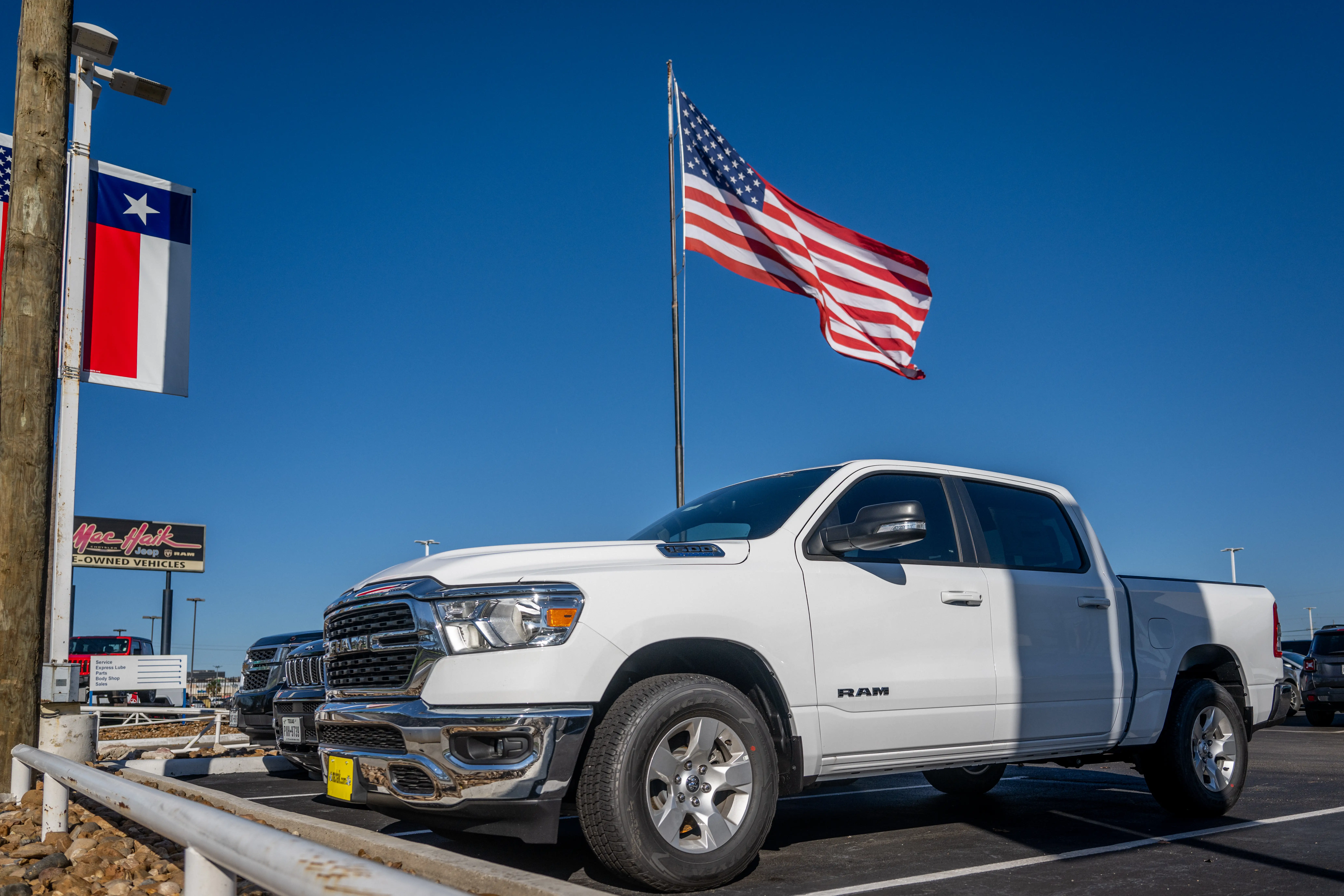 A white Ram 1500 - Stellantis's full-size pickup truck - is parked on a dealership lot in front of an American flag.