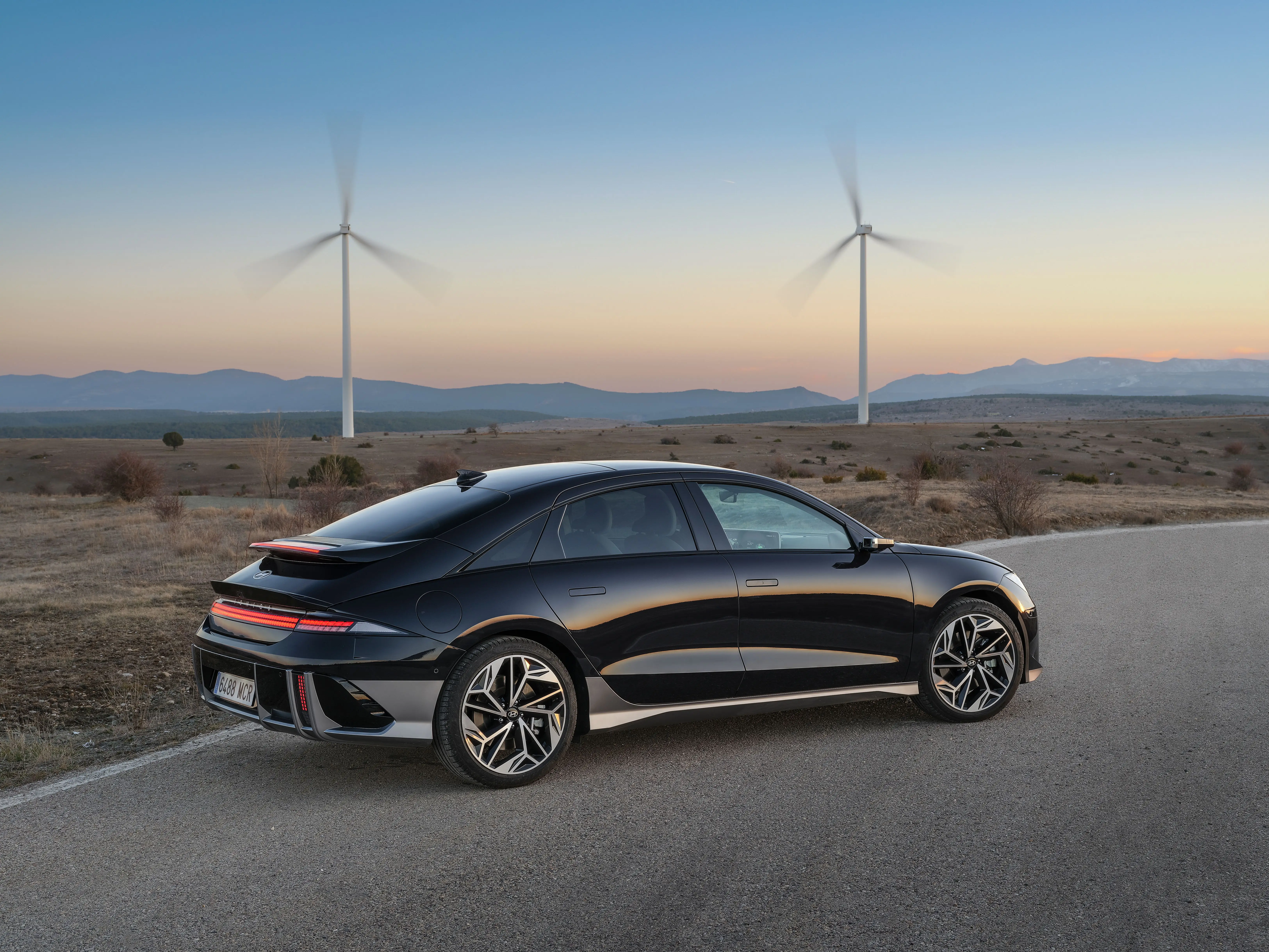 A black Hyundai Ioniq 6 sedan is parked on a section of tar next to a large, sandy field. The sun is setting behind a faraway mountain range, and there are two twirling windmills.