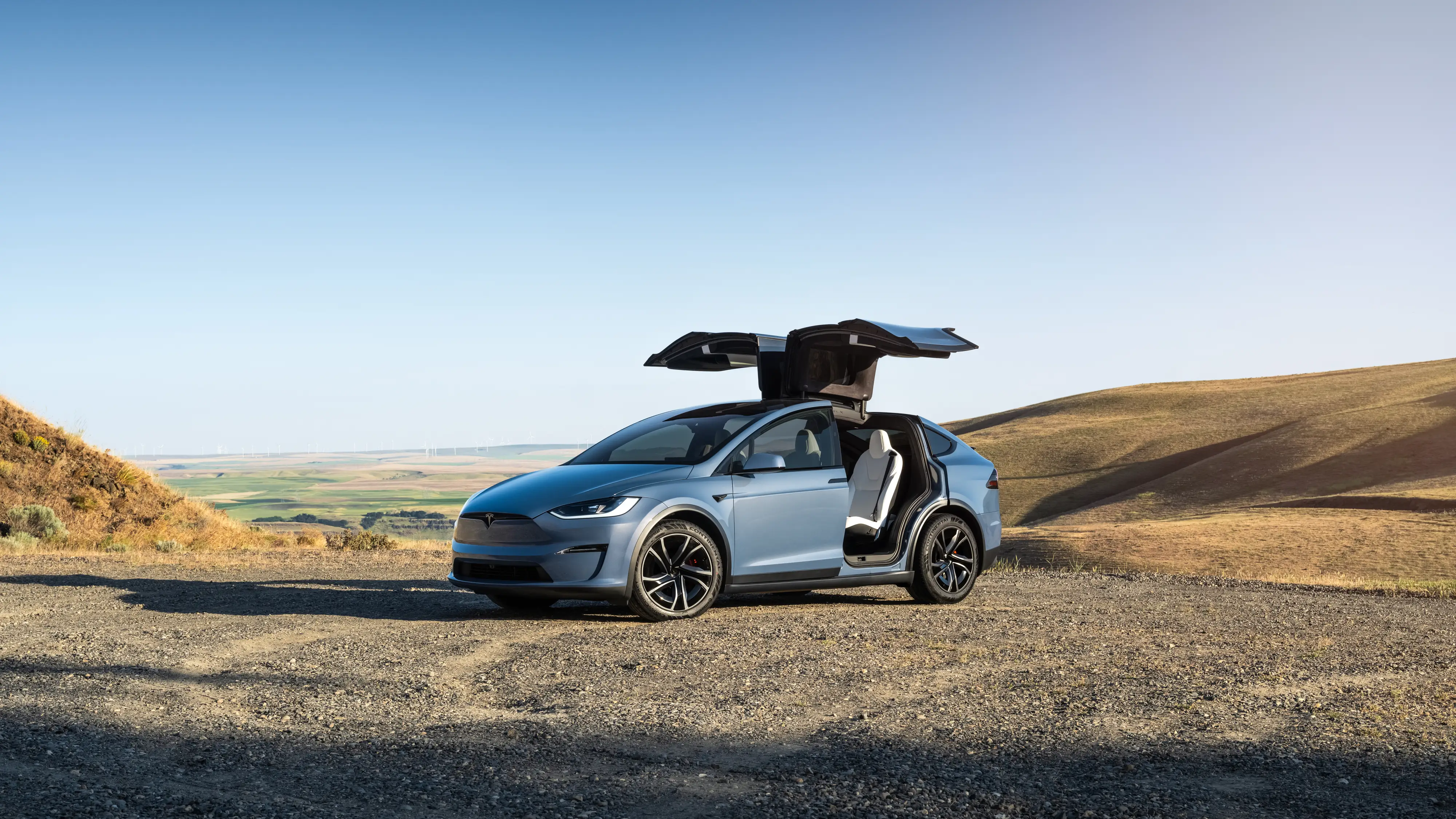 A light blue Tesla Model X is parked on gravel next to a sandy hill. Its gull-wing back doors are open.