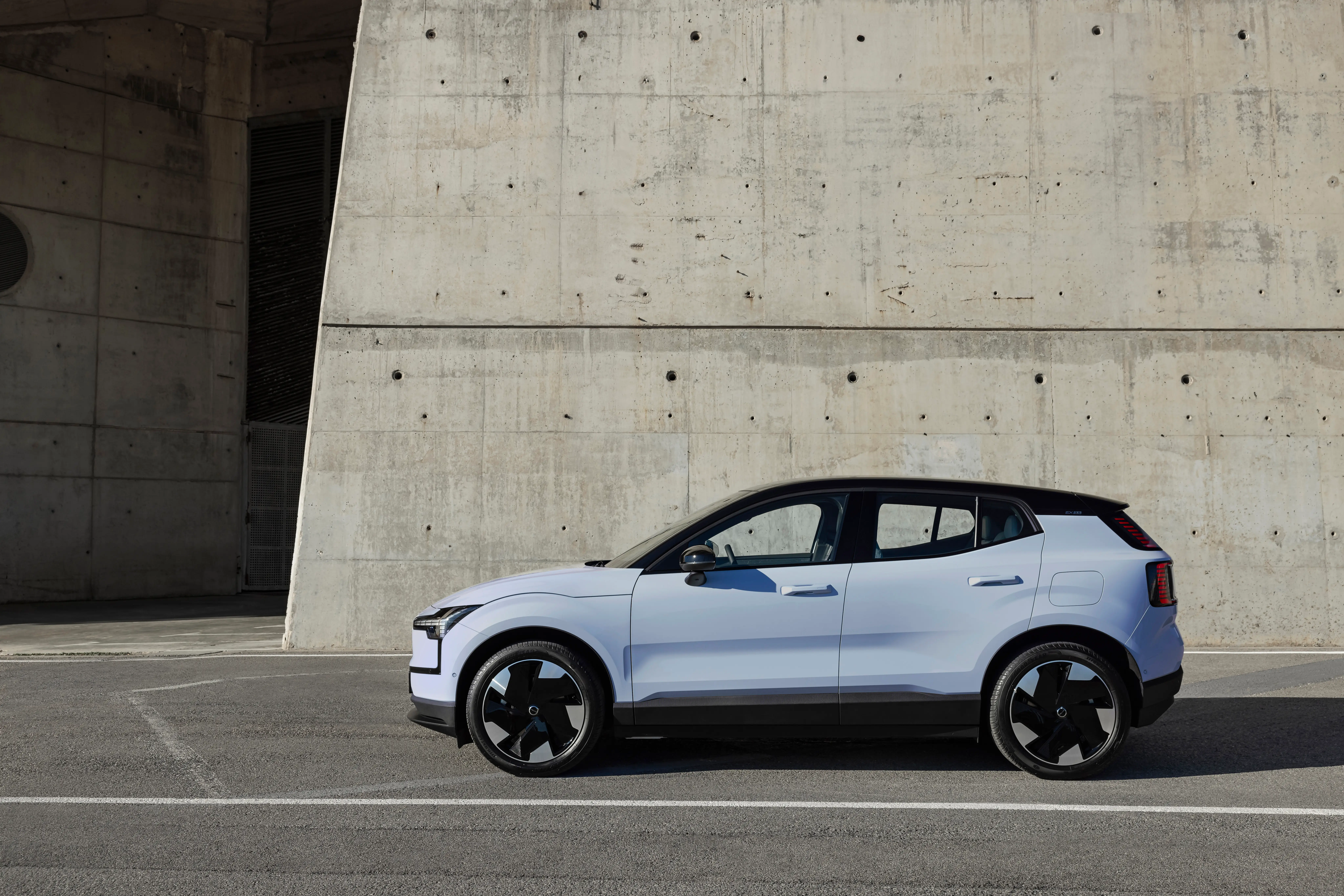 A light blue Volvo EX30 is parked next to a large concrete wall.