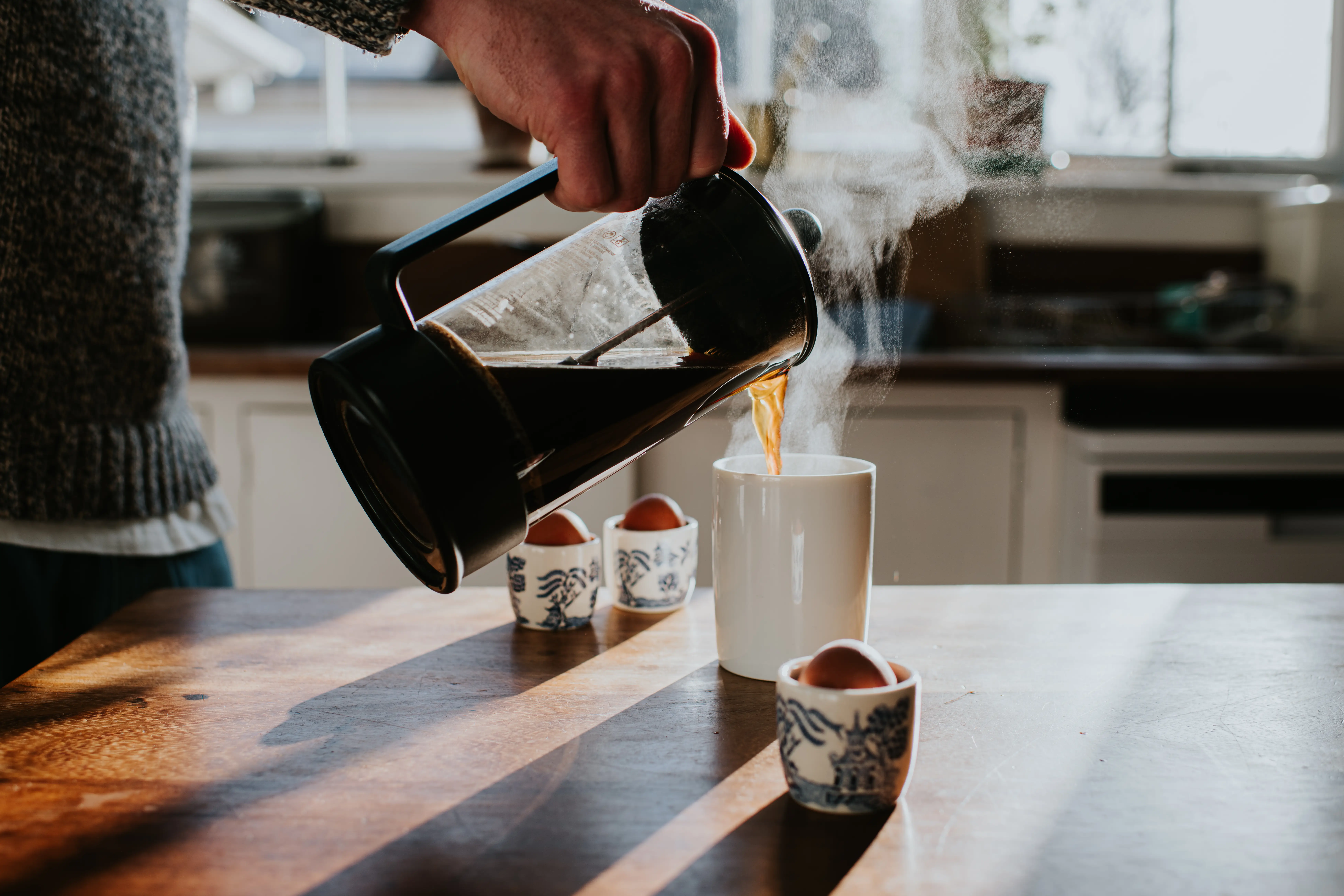 a close up of a hand pouring a french press coffee into a mug with steam rising in a kitchen lit by early morning sunlight