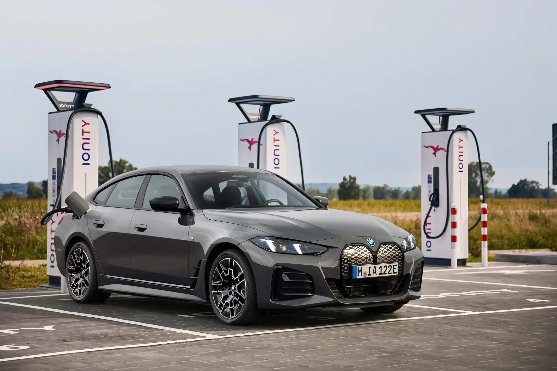 A grey BMW i4 charges at a public charging station next to a grassy field.