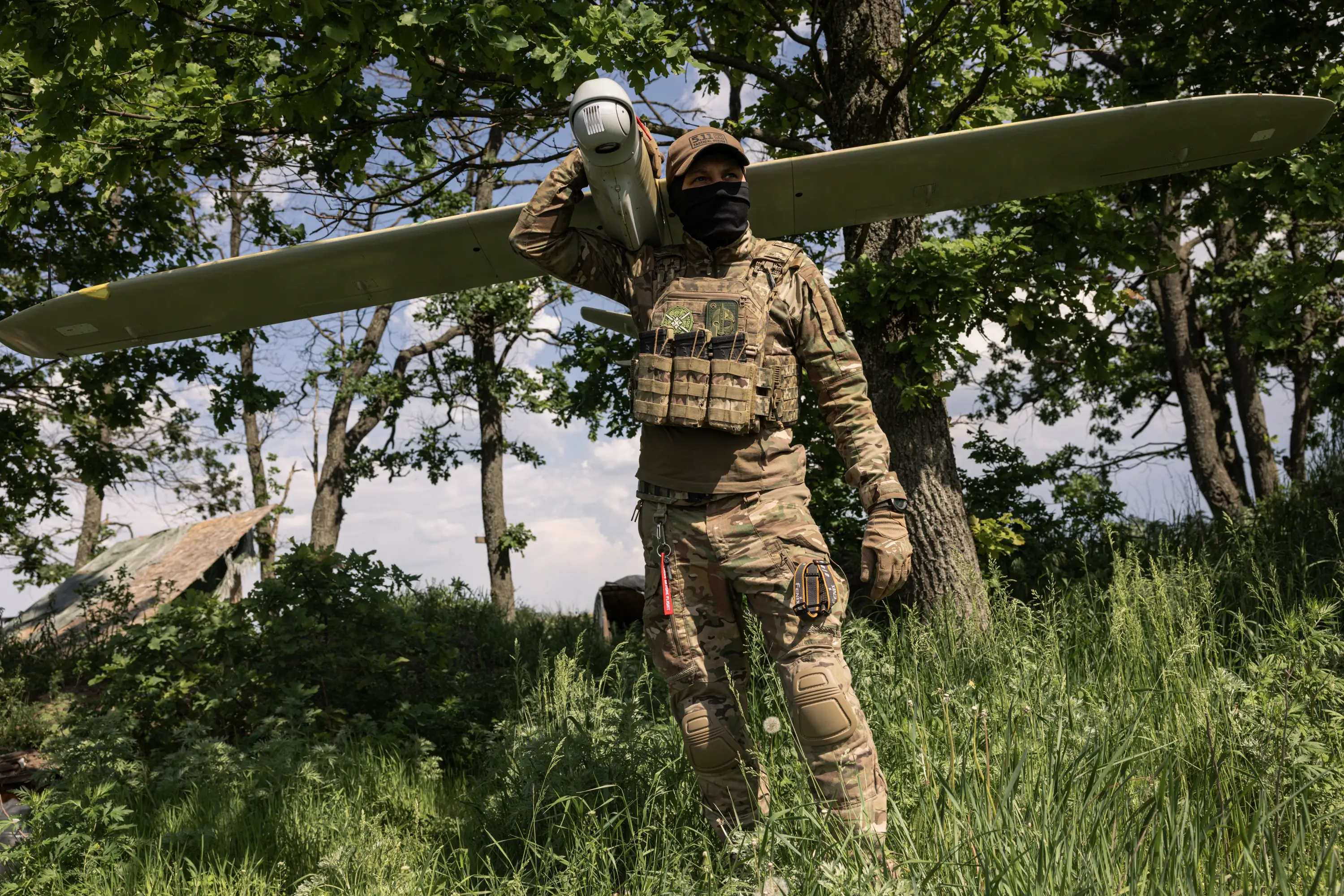 A man in camouflage gear stands in a field with trees behind him with a large winged drone on his shoulder