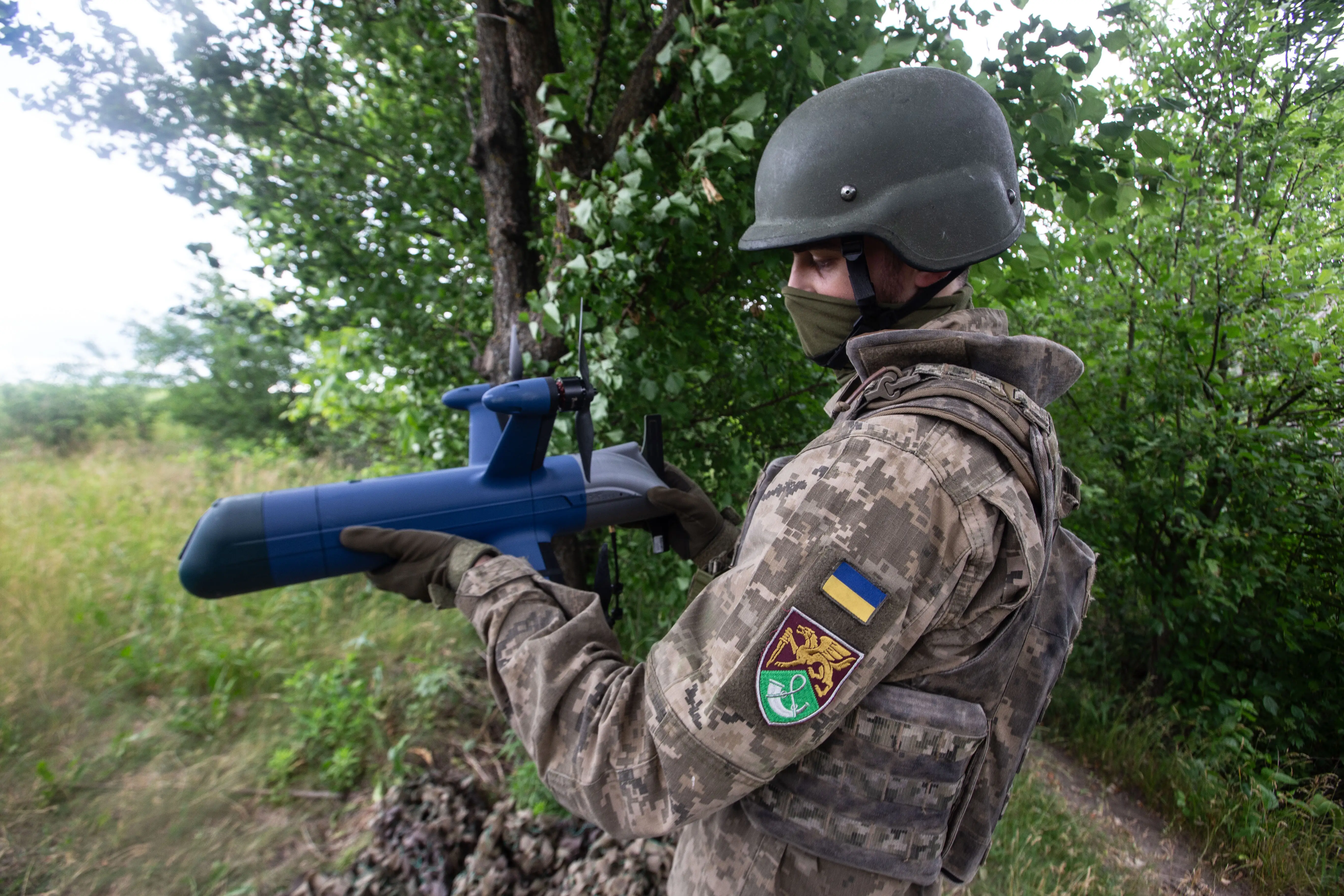 A man in camouflage gear and a helmet stands holding a blue drone among green trees and bushes