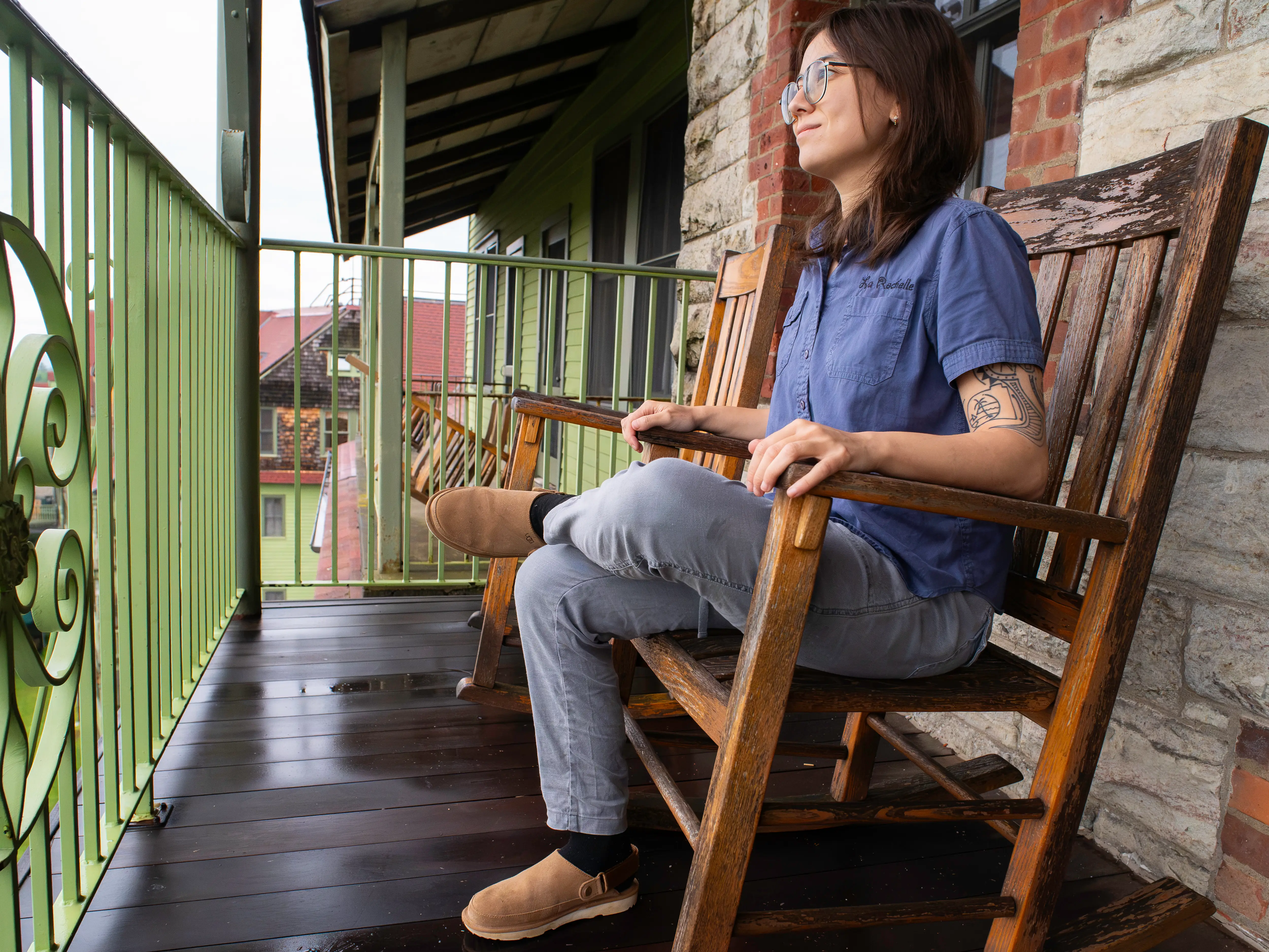 Person wearing glasses sits in a wooden rocking chair on a covered porch with green railing at Mohonk Mountain House