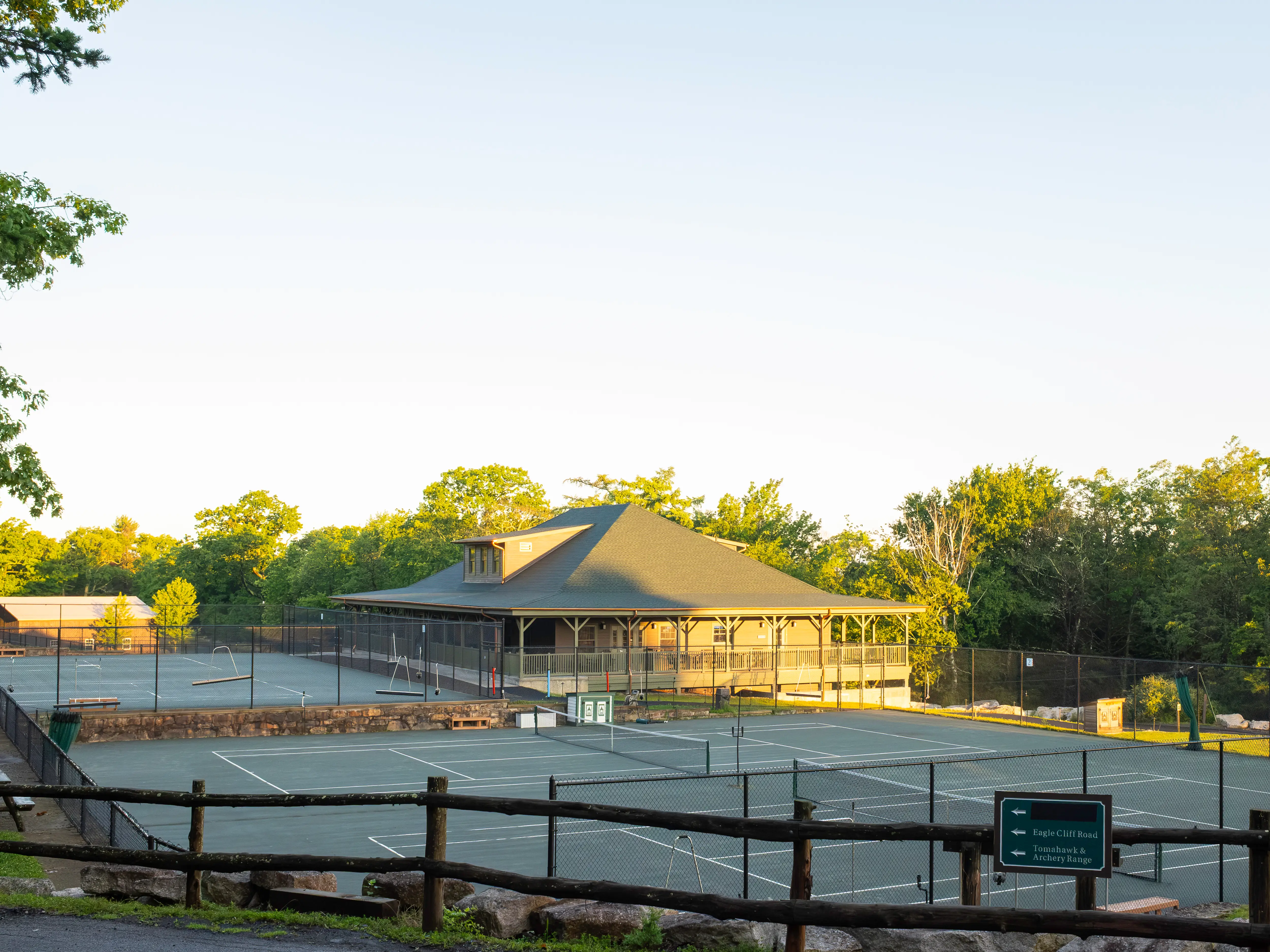 Tennis courts and a clubhouse sit among trees in warm late-day sunlight.
