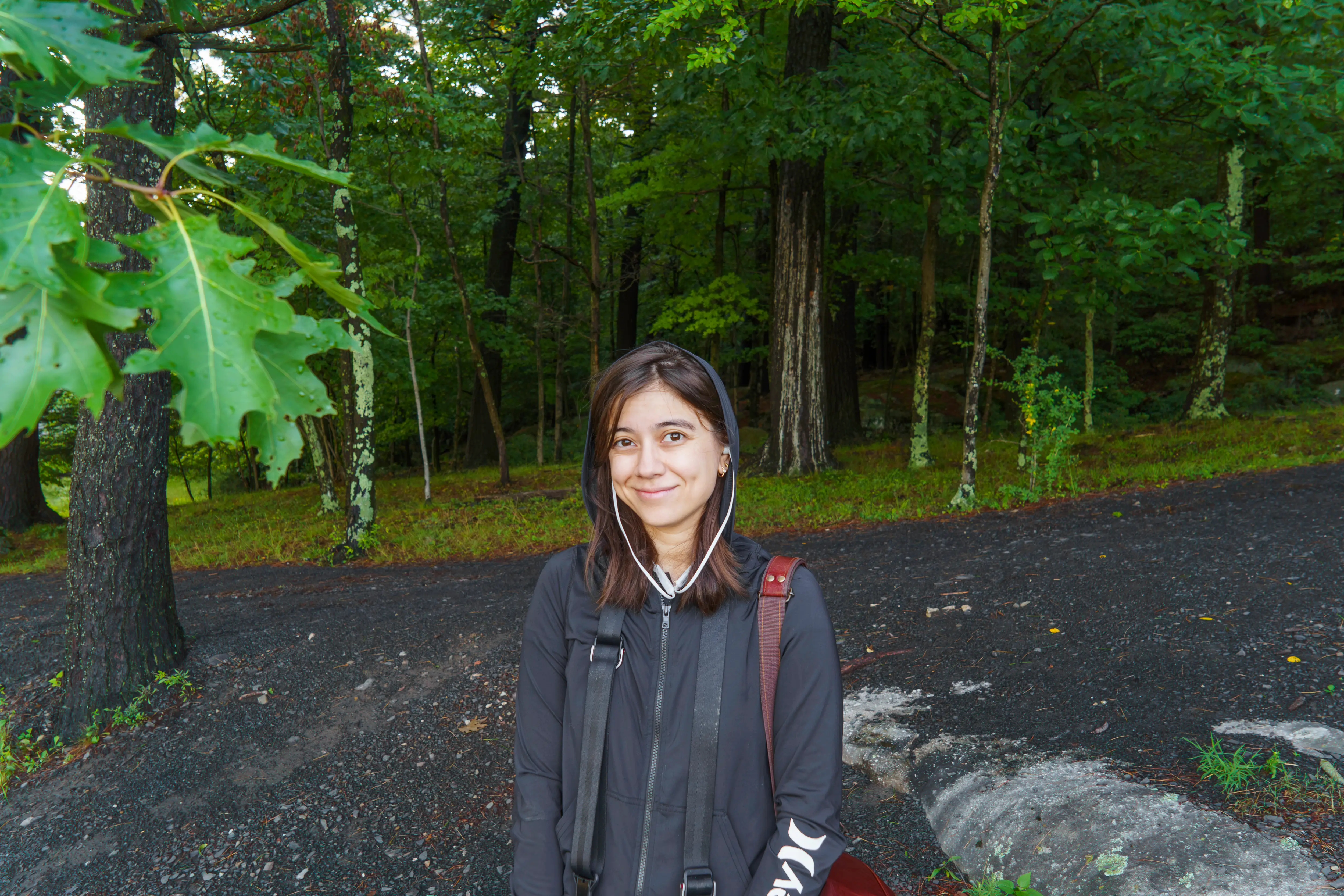Person wearing a hooded jacket and earbuds stands on a wooded path surrounded by green forest.