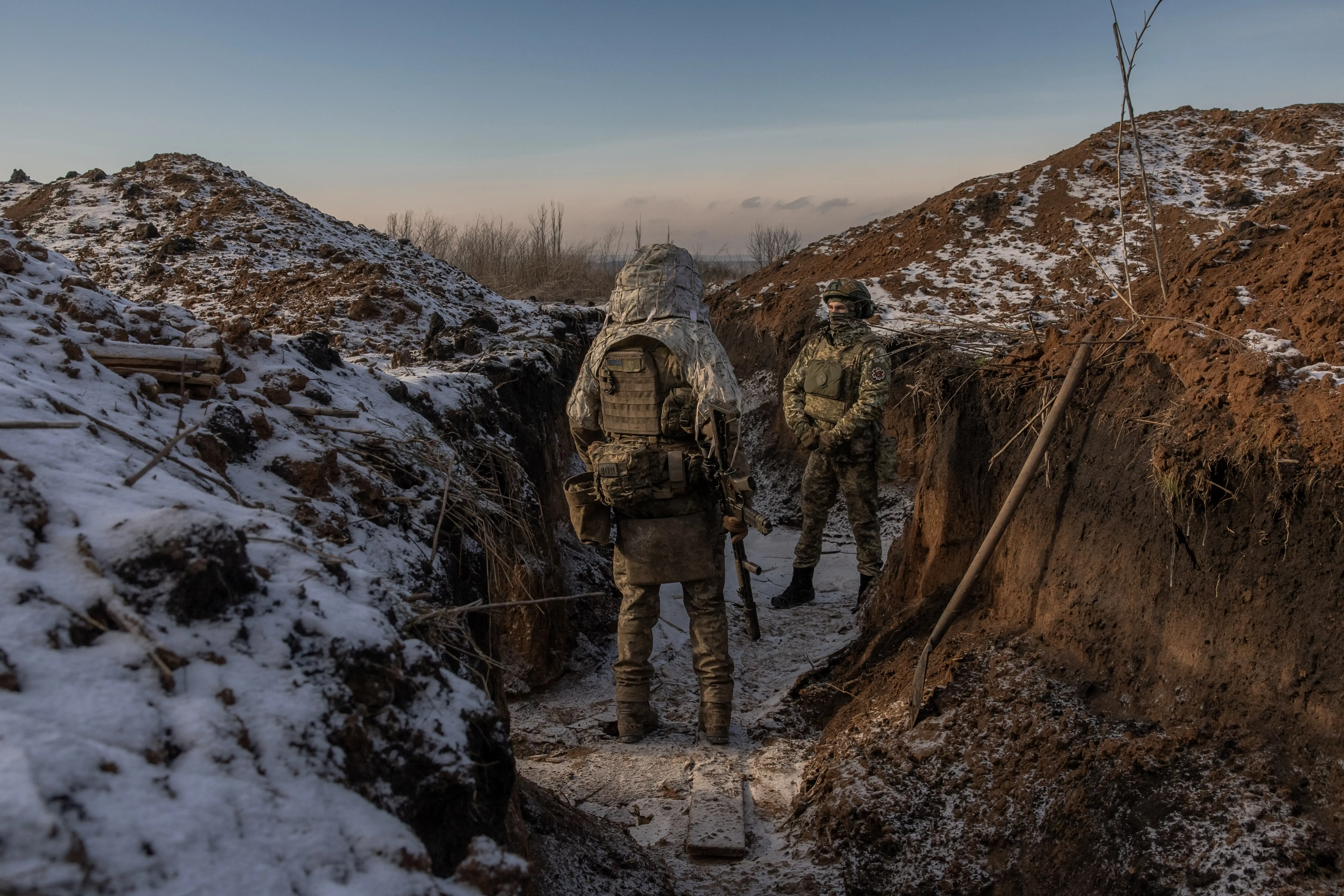 Two camouflaged military personnel stand in a snow-dusted trench between earthen embankments at dusk.
