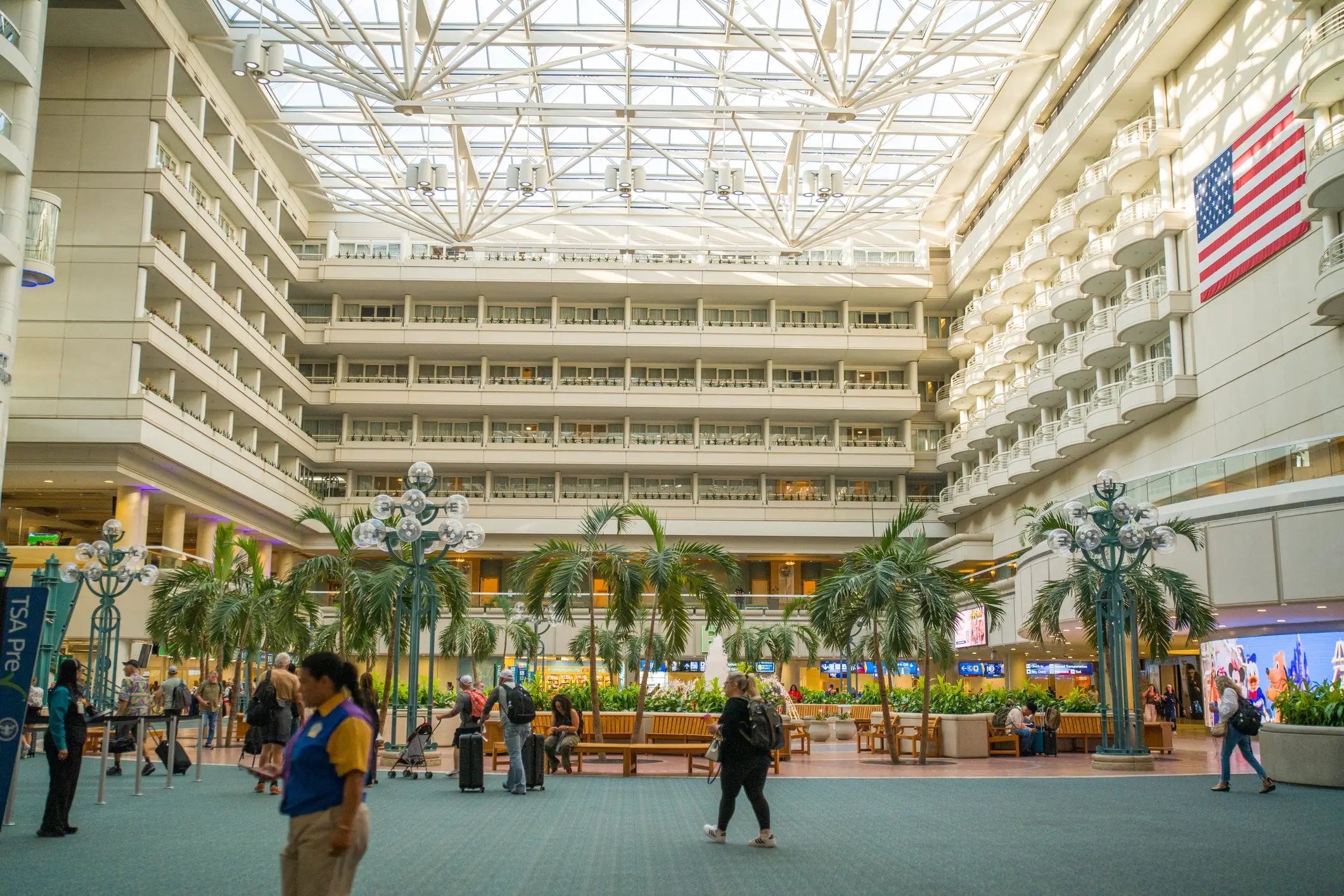 Travelers move through Orlando International Airport with palm trees, balconies, skylight, and a large U.S. flag.